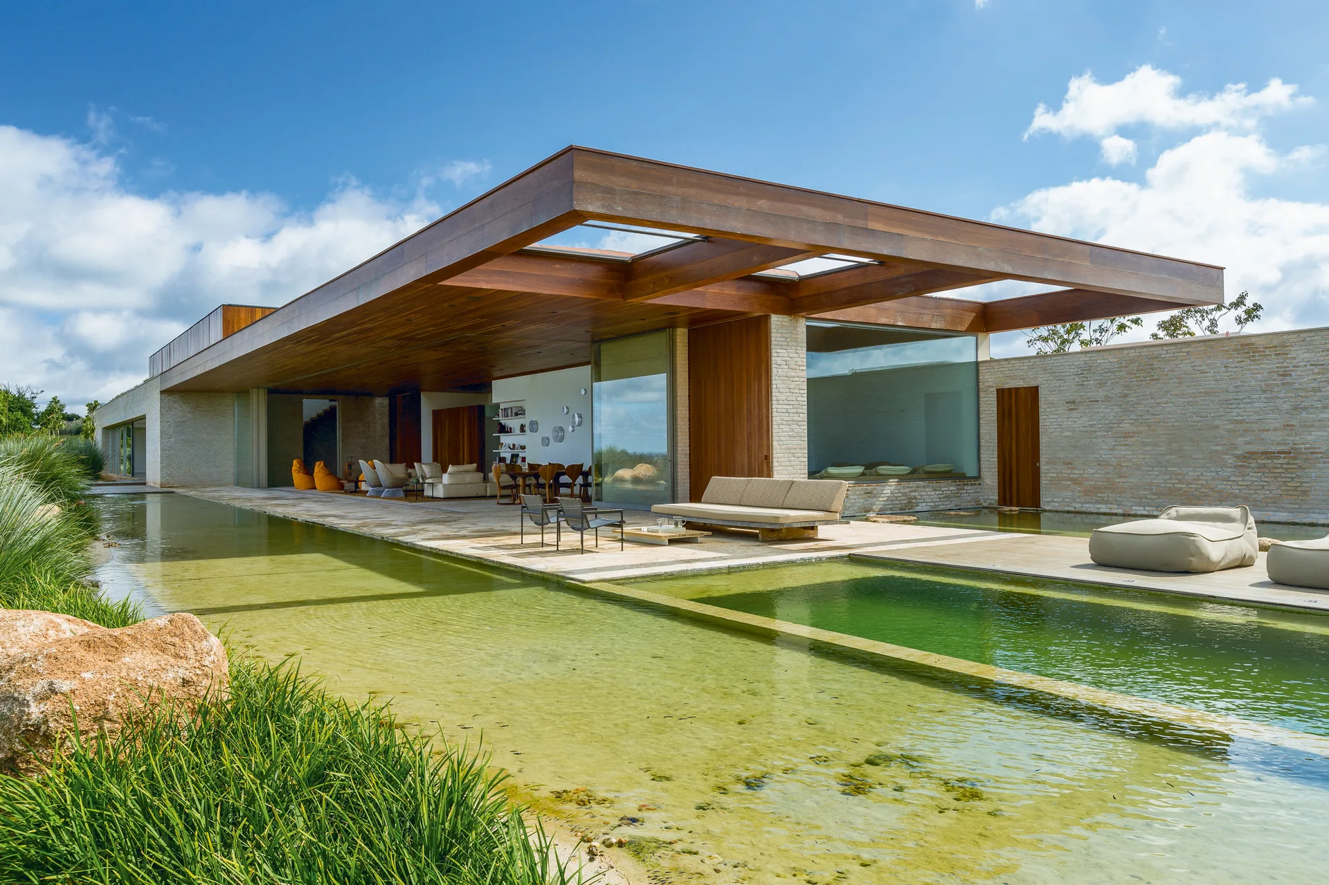 A modern outdoor patio features a large swimming pool, a massive cantilevered wood ceiling overhang, stone walls, and minimalist seating.