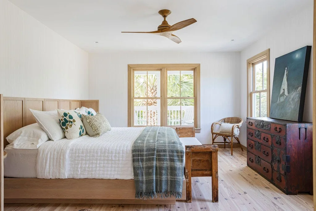 A bright bedroom featuring a light wood bed, white vertical paneled walls, a rustic dark wood chest of drawers, and a wooden ceiling fan.