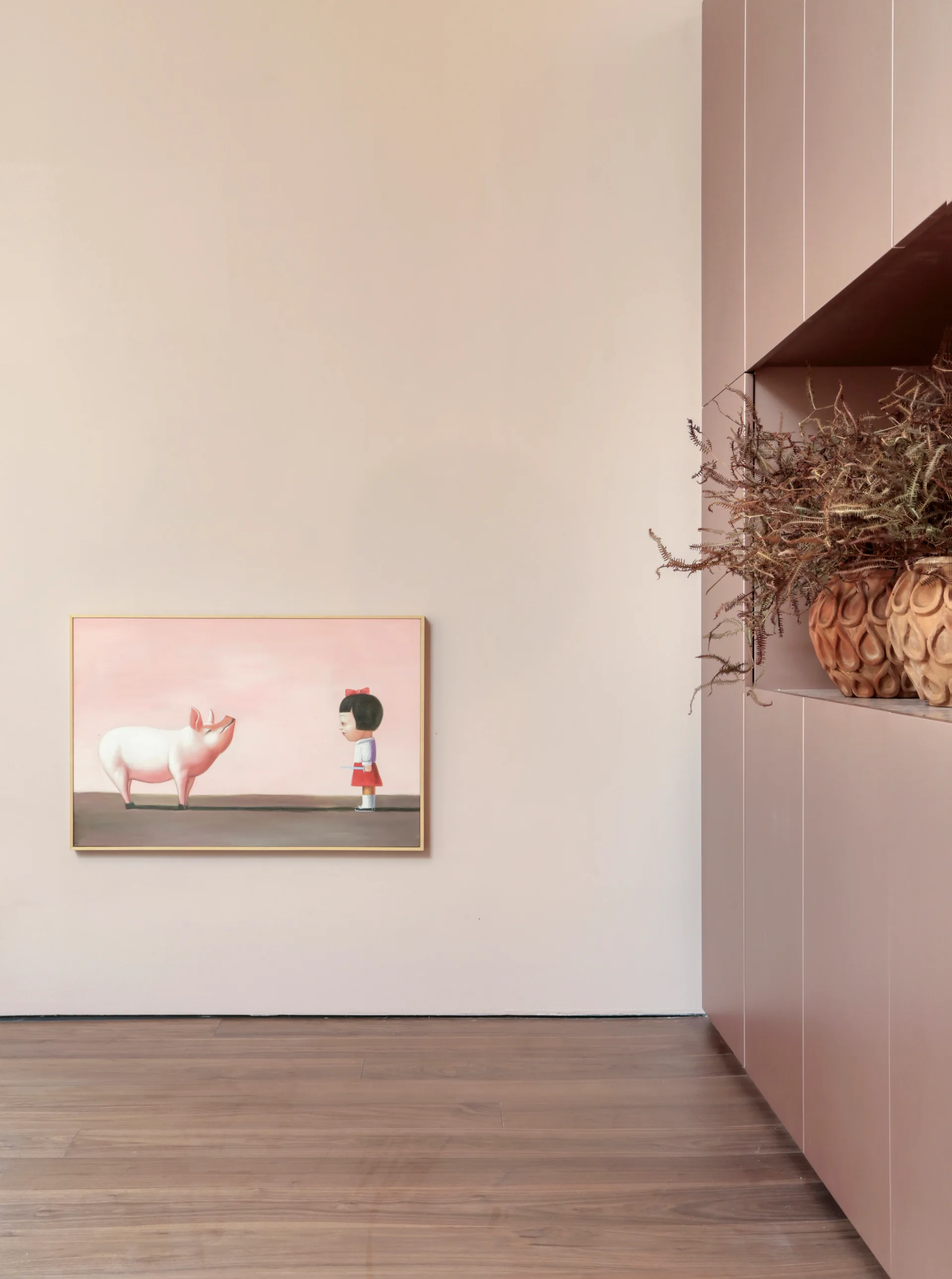 A minimalist room with dusty pink walls, built-in flush cabinetry, warm hardwood floors, and a low-hung painting of a girl and a pig.