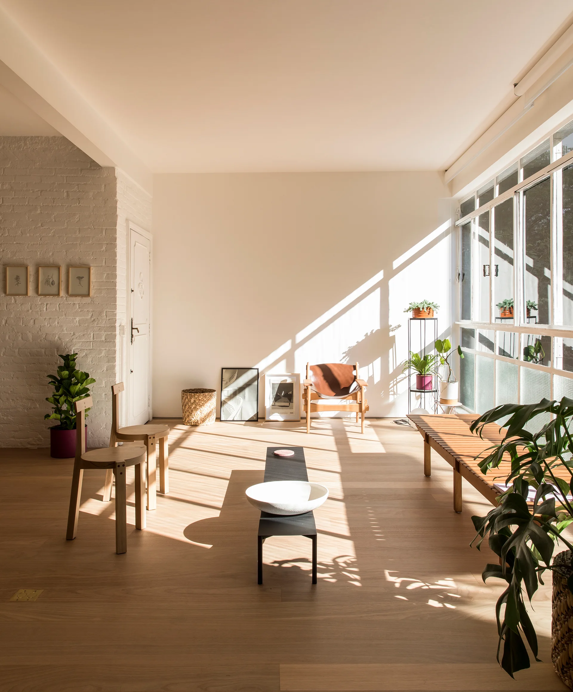 A minimalist living room filled with bright sunlight features a slatted wooden bench, wooden chairs, and leaning art frames on light oak floors.