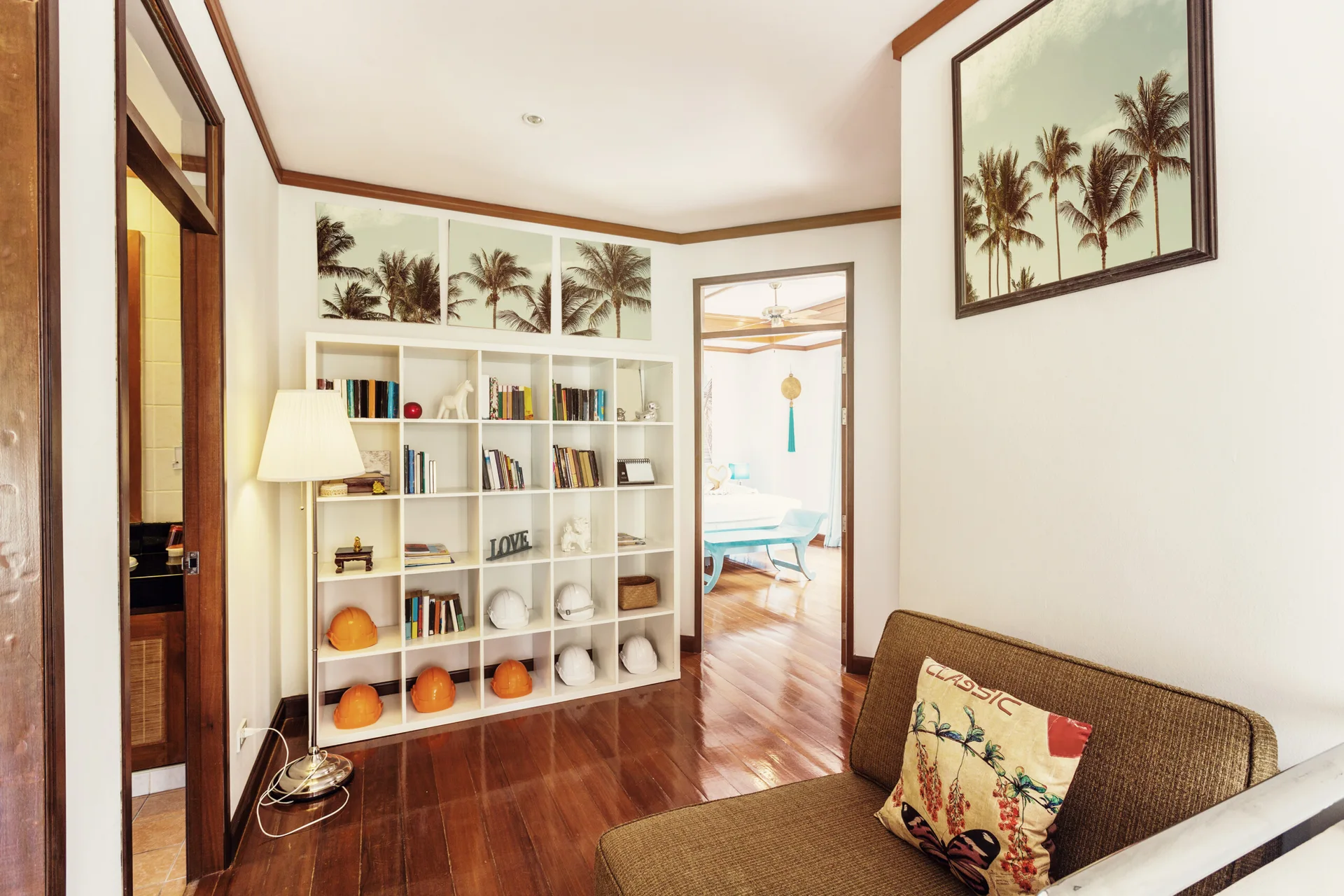 A transitional hallway features a white grid bookshelf, a brown sofa, and framed palm tree art on polished hardwood floors.