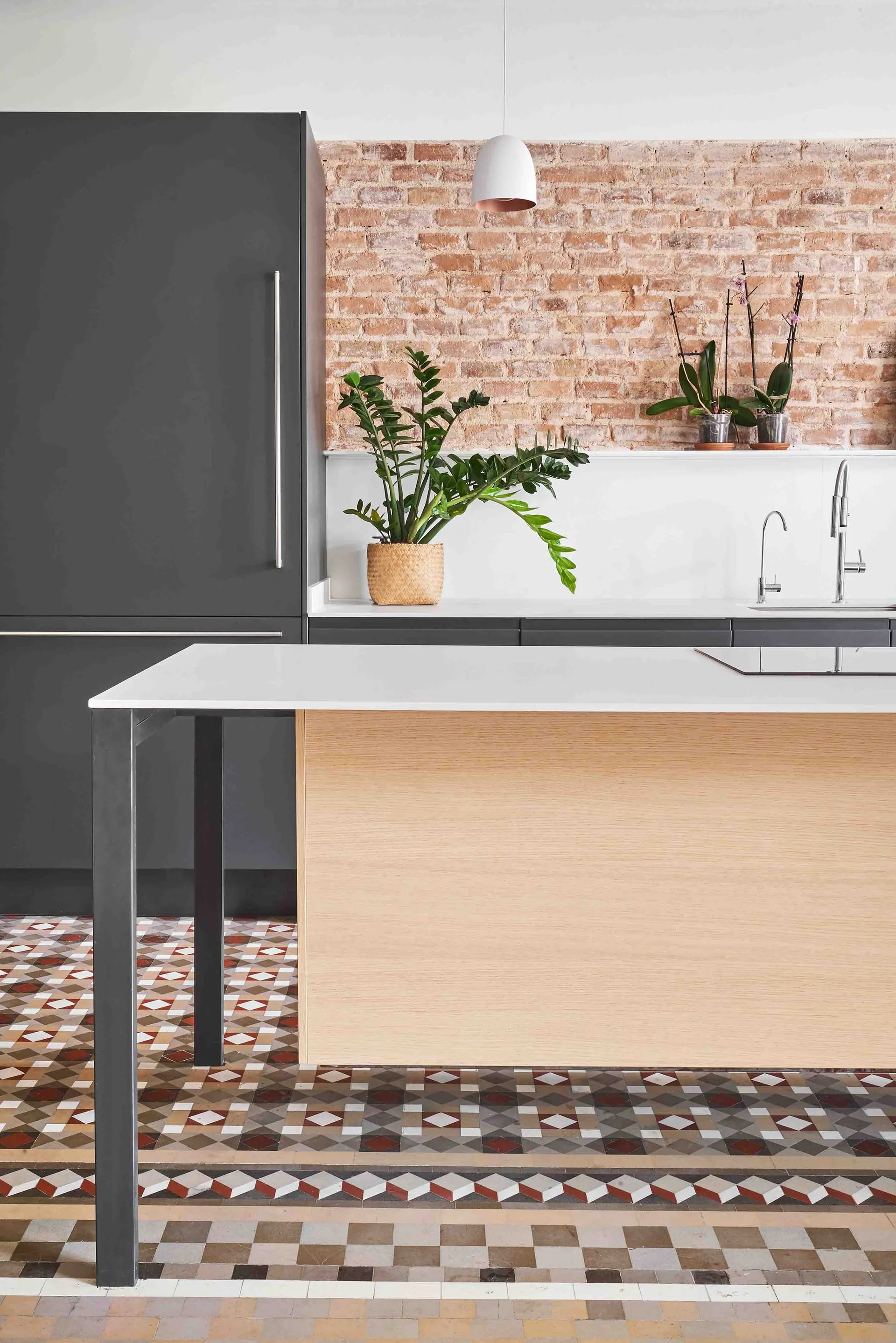 A modern kitchen features a wood-fronted island, matte grey cabinetry, an exposed brick backsplash, and geometric tiled flooring.