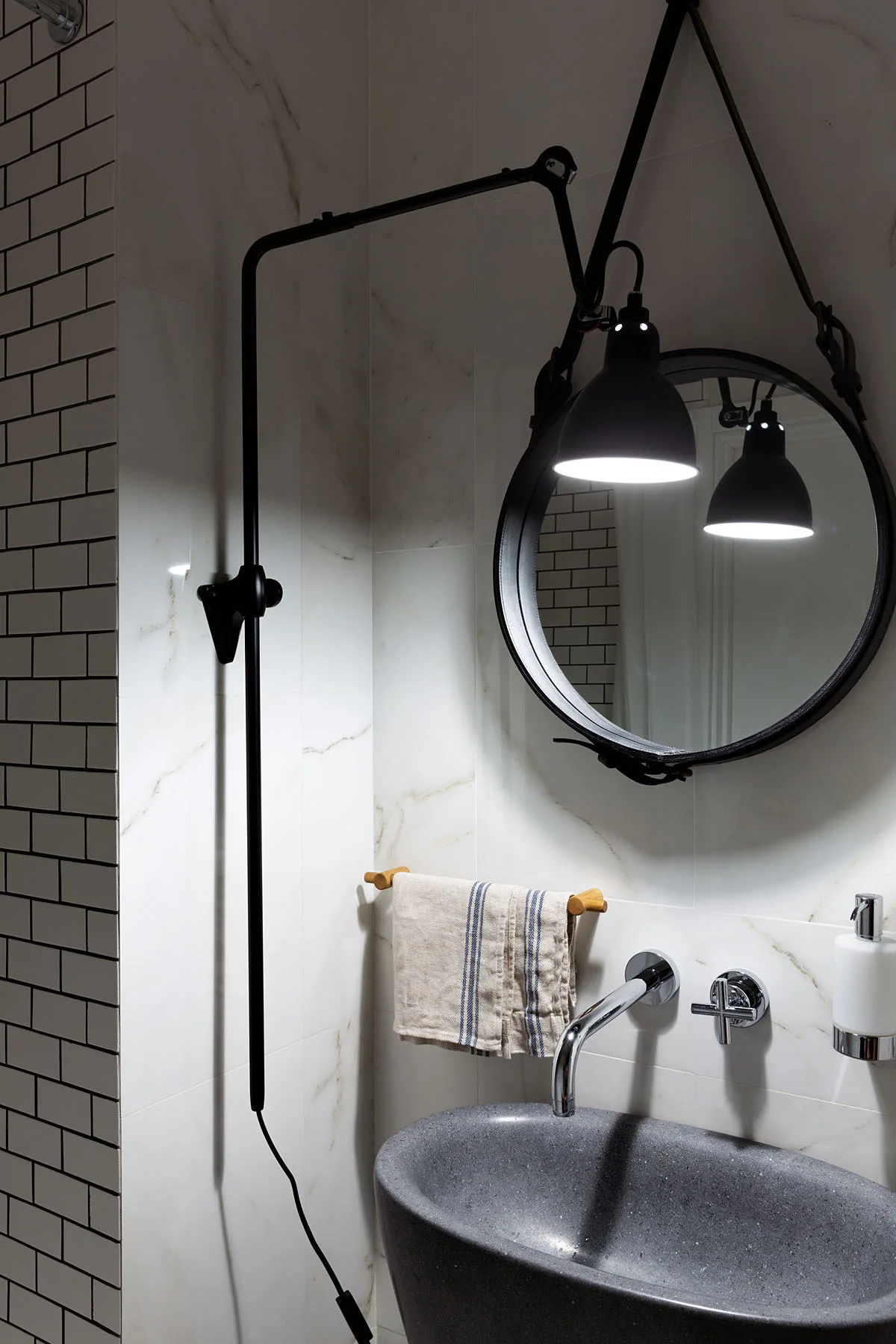 A grey stone basin sink sits beneath a wall-mounted chrome faucet, a circular mirror, and an adjustable black swing-arm lamp against marble tiles.