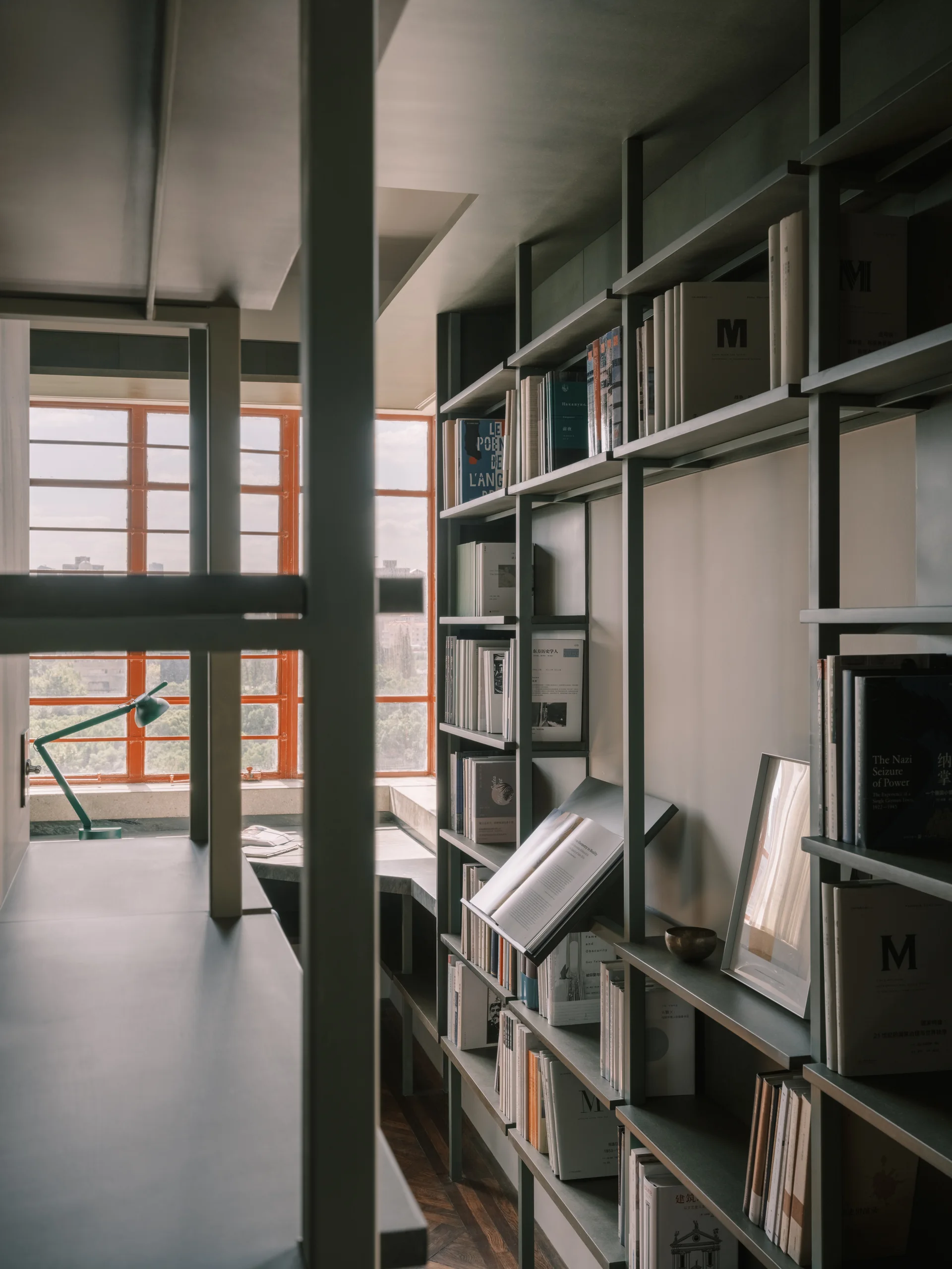 Floor-to-ceiling gray metal bookshelves stand next to a desk positioned before a large grid window with a bright orange frame.