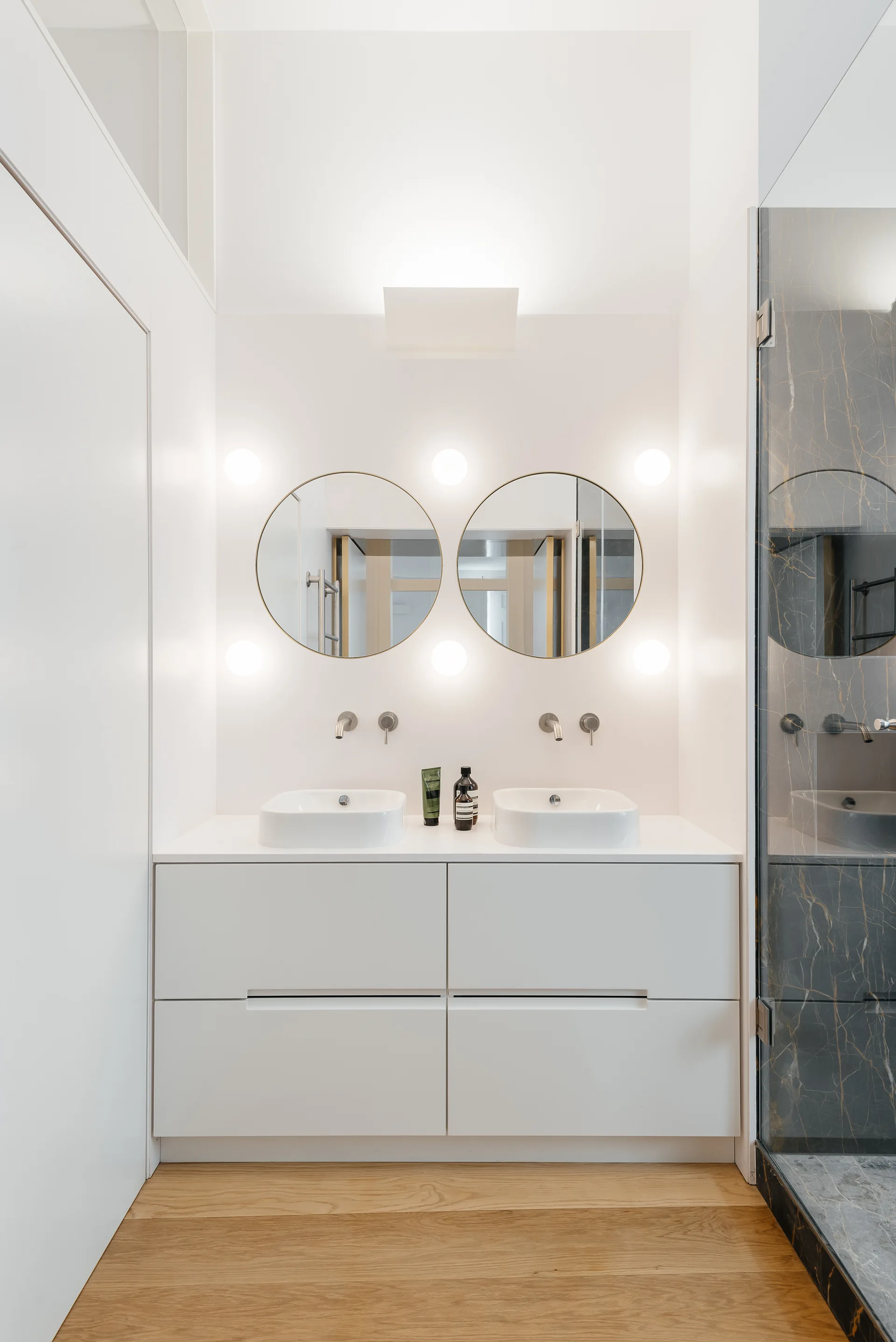 A modern bathroom vanity setup with two white vessel sinks, circular mirrors, round sconce lighting, and a glass shower enclosure.