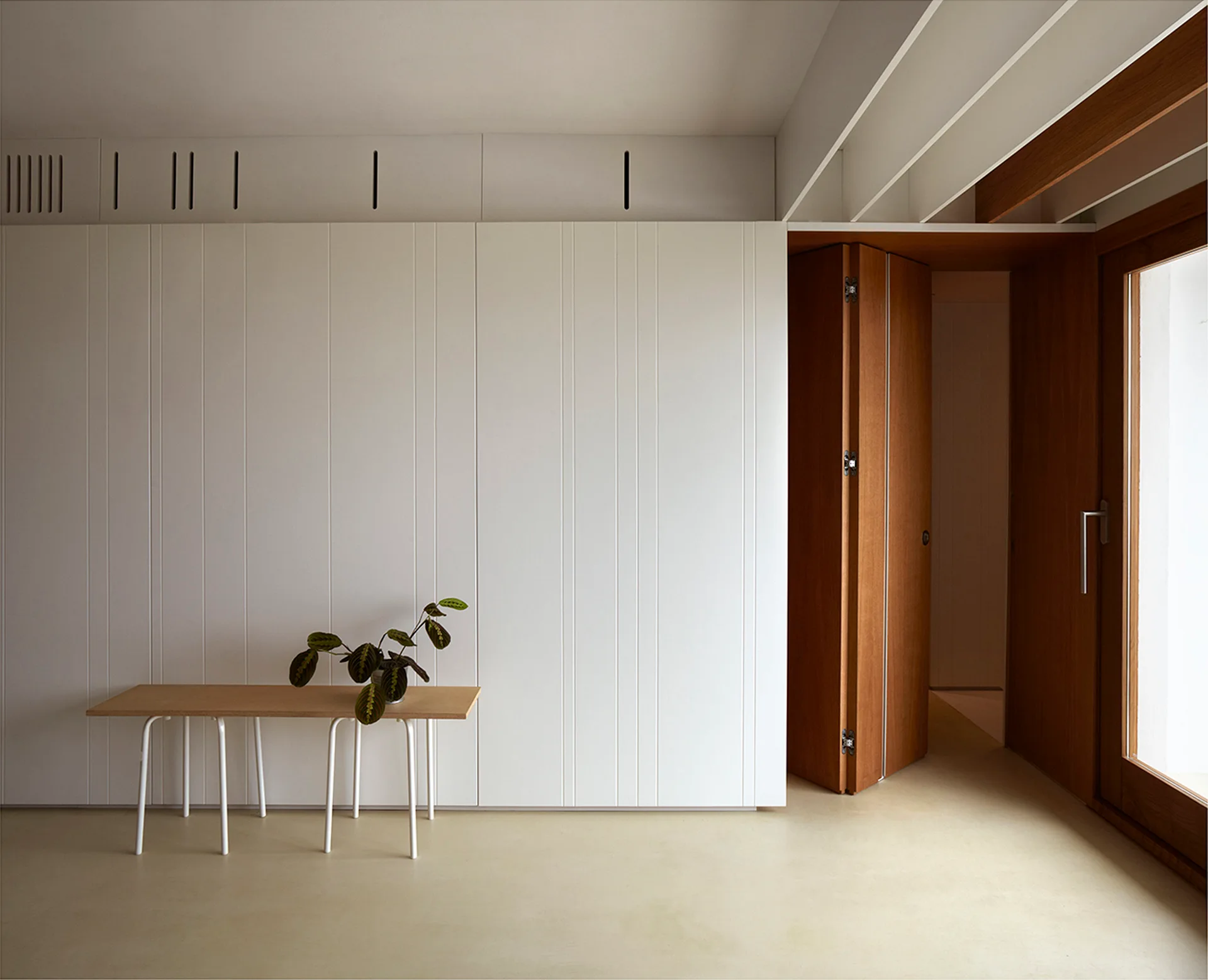 A minimalist entryway features a light wood bench with white legs beneath a tall, vertically paneled white wall next to a warm wood folding door.