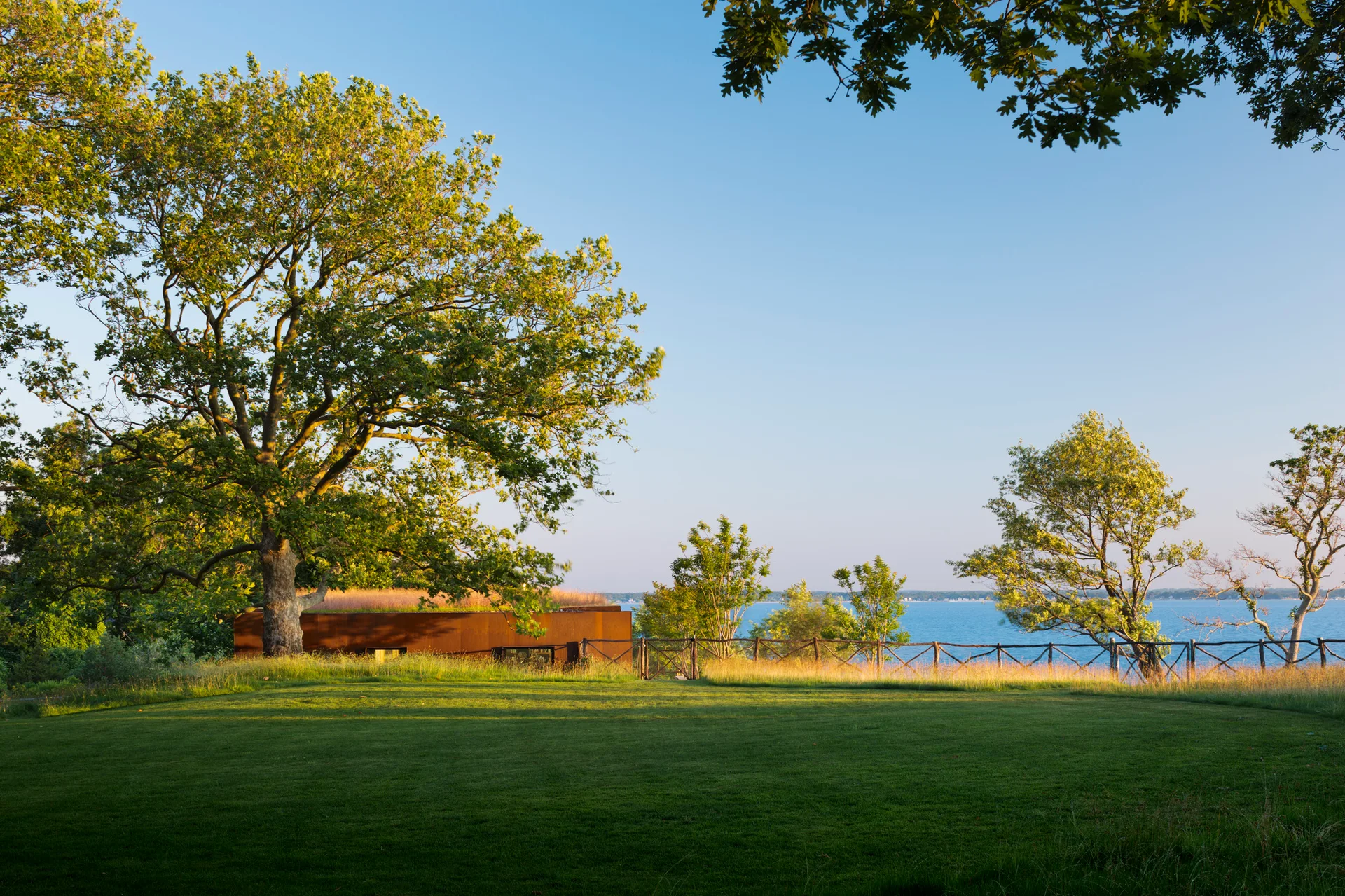 A wide lawn and rustic fence overlook the ocean, with a modern rust-colored house featuring a green roof settled into the grassy landscape.