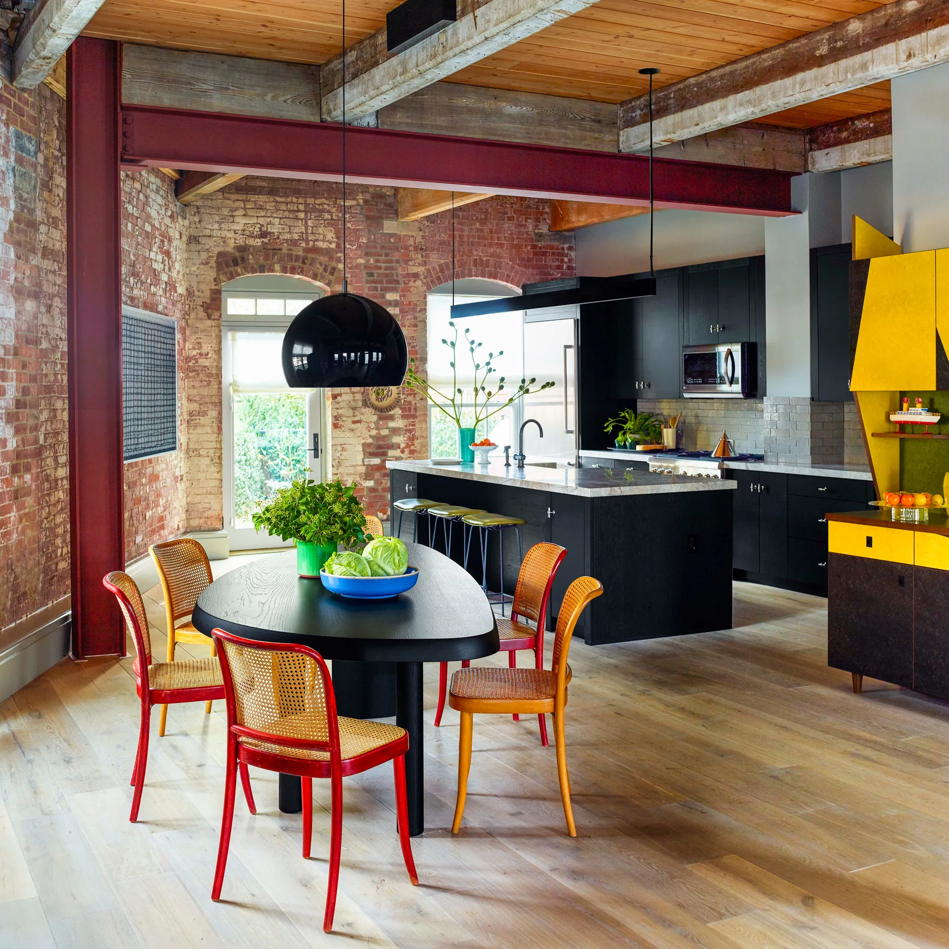 An industrial loft kitchen and dining area features exposed brick walls, a black oval dining table, colorful cane chairs, and a black kitchen island.