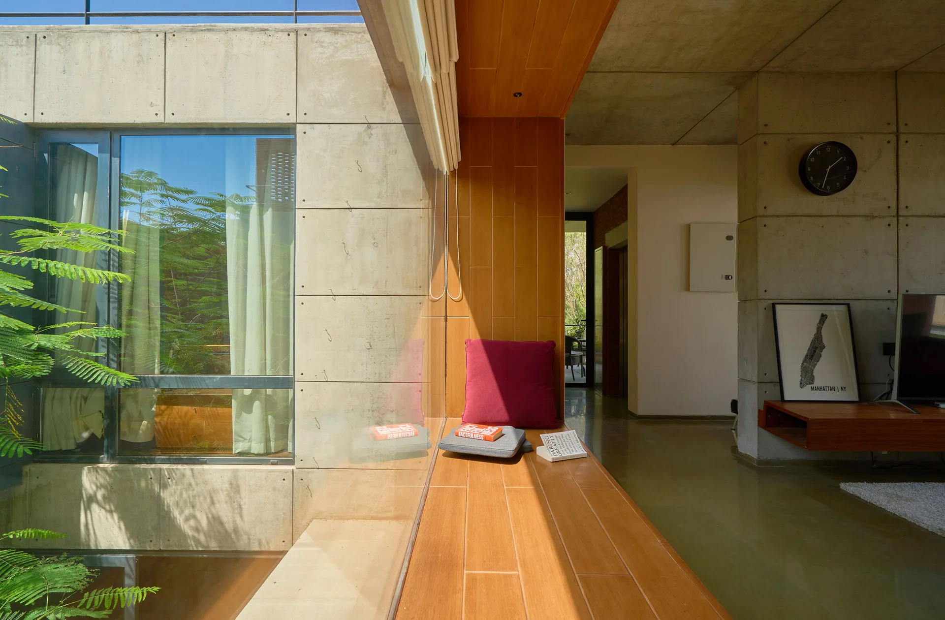 A long, wooden built-in window bench with a single burgundy cushion is set against a large window and exposed concrete panel walls.