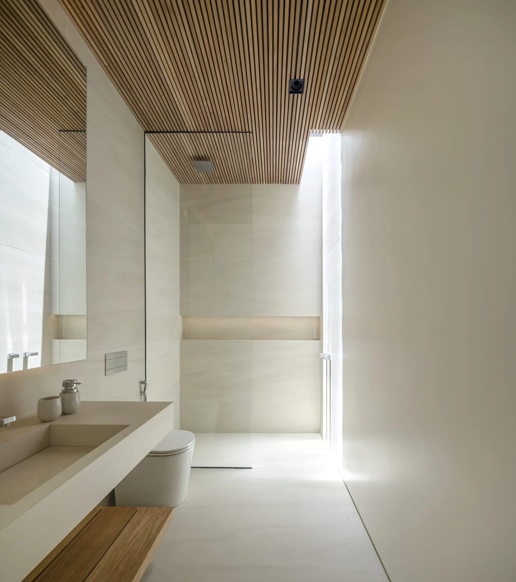 A minimalist bathroom features a warm wood-slat ceiling, an integrated stone vanity sink, a glass shower partition, and cream-colored walls.