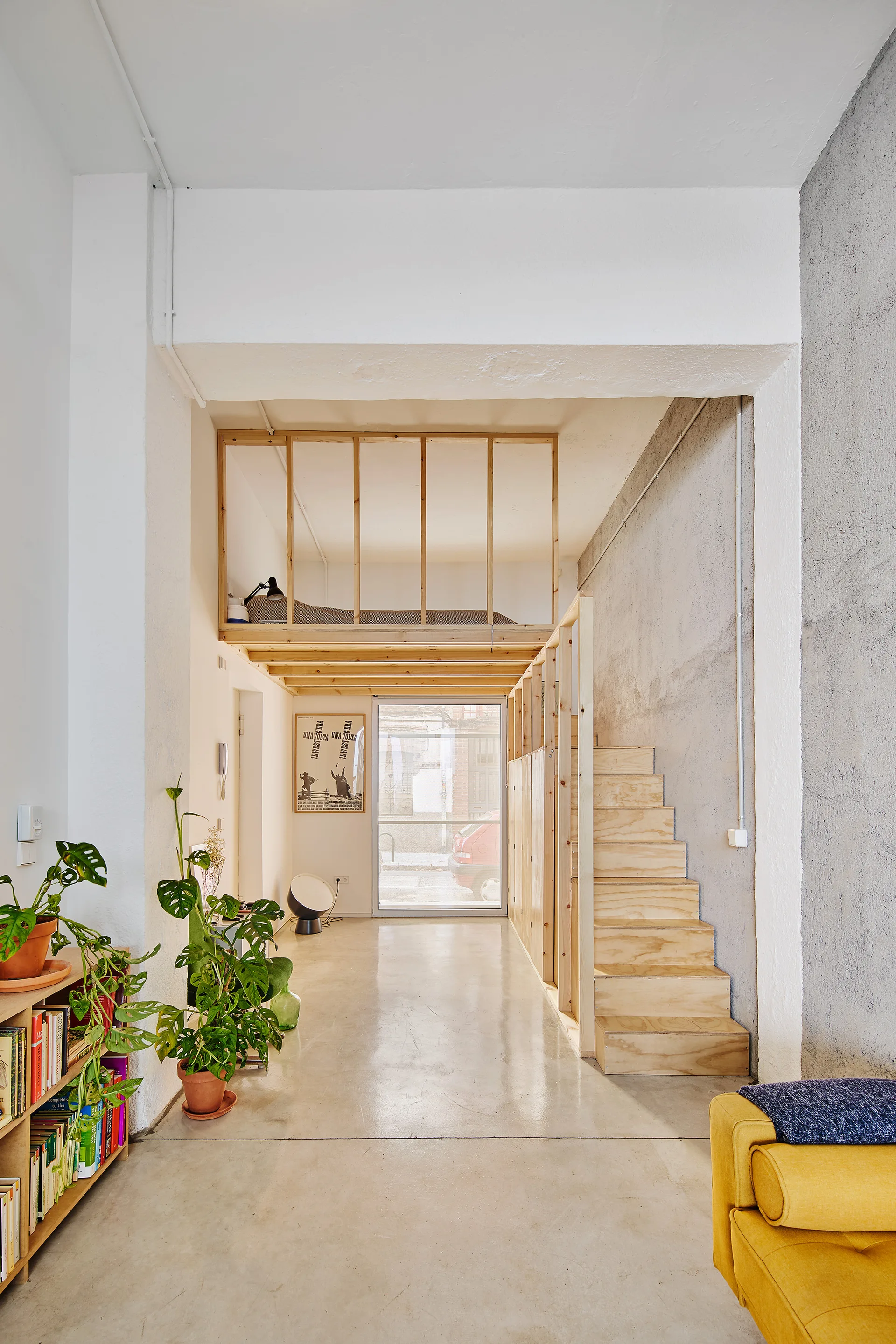 A light-filled minimalist room features a custom plywood loft structure with a built-in staircase next to a textured concrete wall.