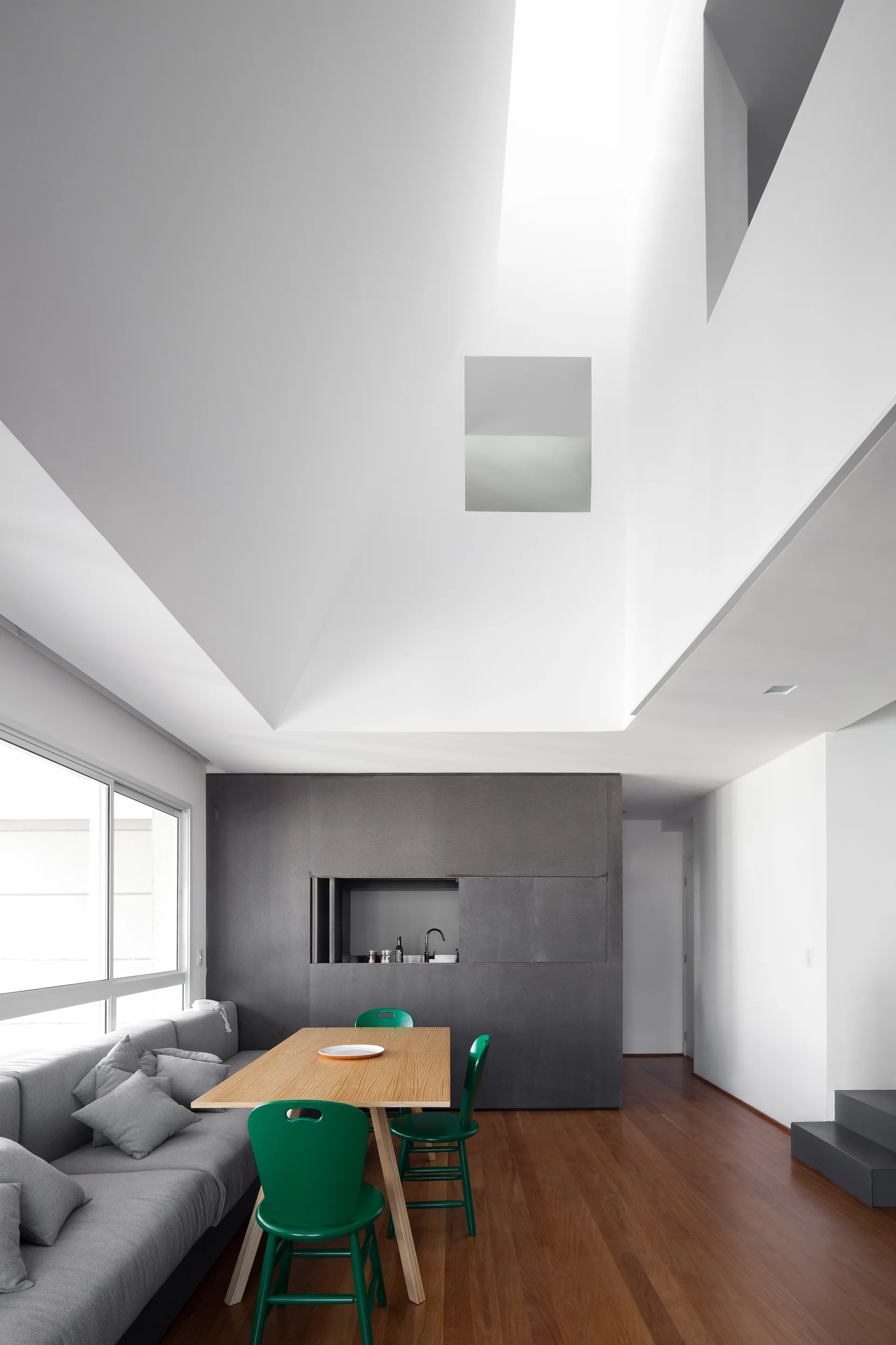 A bright modern dining room with a wooden table, emerald green chairs, and a long gray bench under an angular skylight.