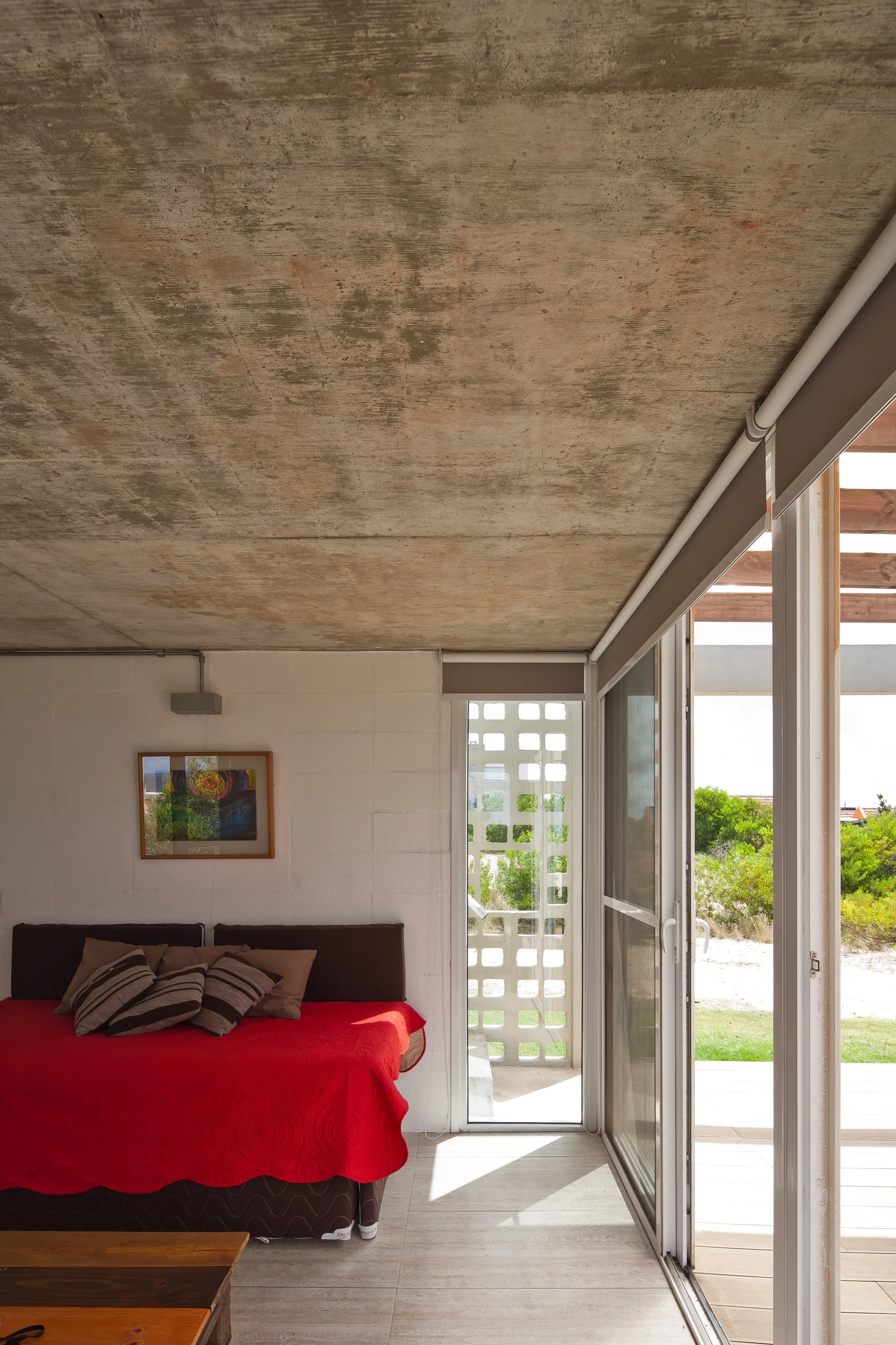 A bedroom features a daybed with a red quilt against a white brick wall, under an exposed concrete ceiling with sliding glass doors opening to a patio.