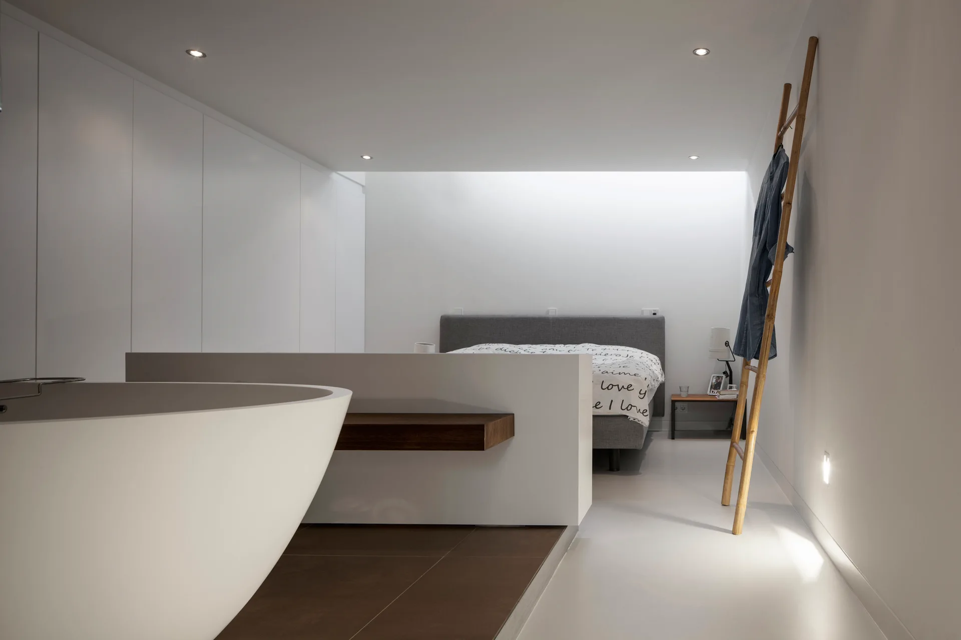 A minimalist open-concept bedroom and bathroom featuring a white freestanding bathtub in the foreground and a grey bed behind a low partition wall.