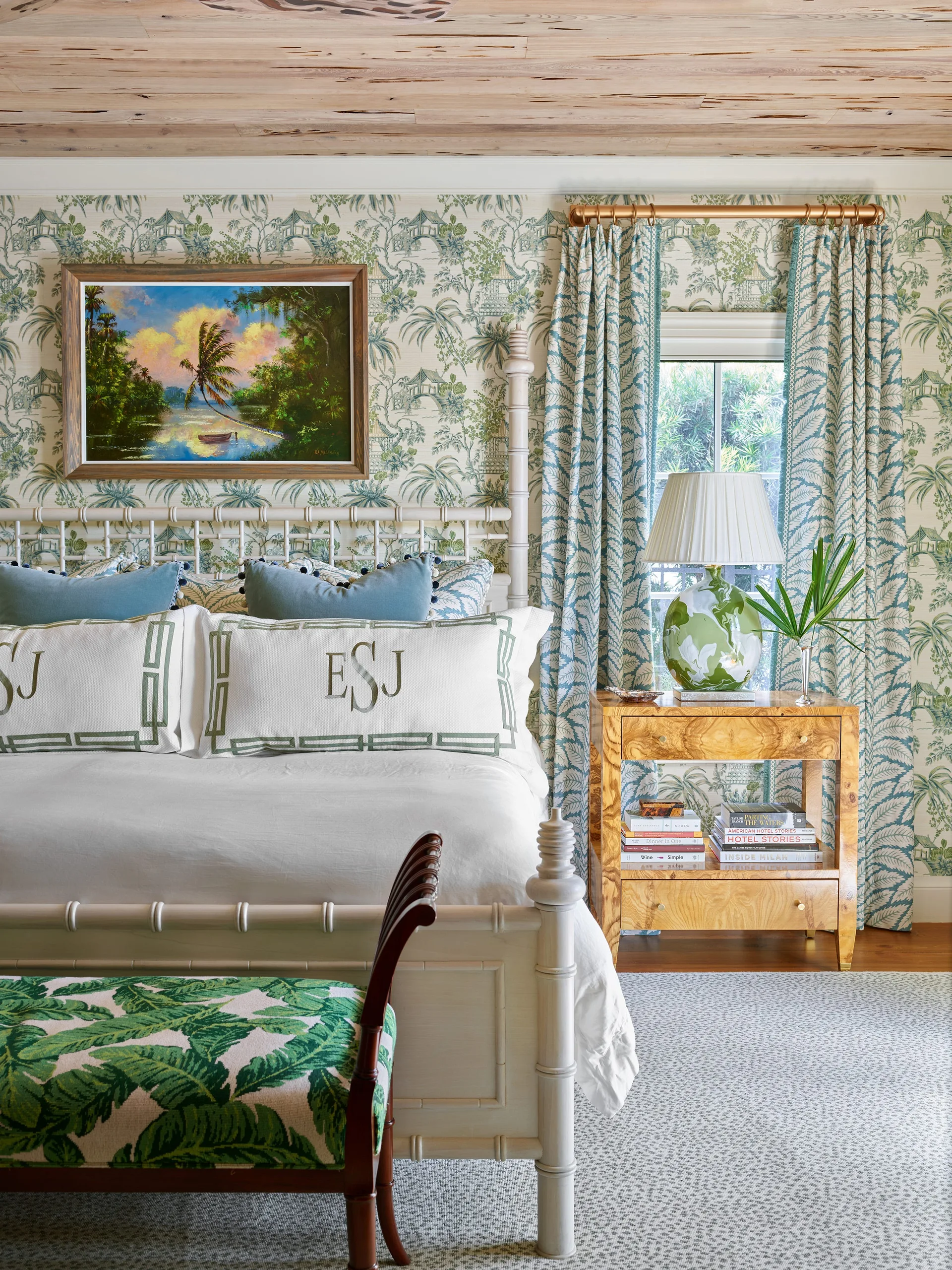 A bedroom featuring a white spindle bed with monogrammed pillows, green chinoiserie wallpaper, and a burl wood nightstand beneath a wood plank ceiling.