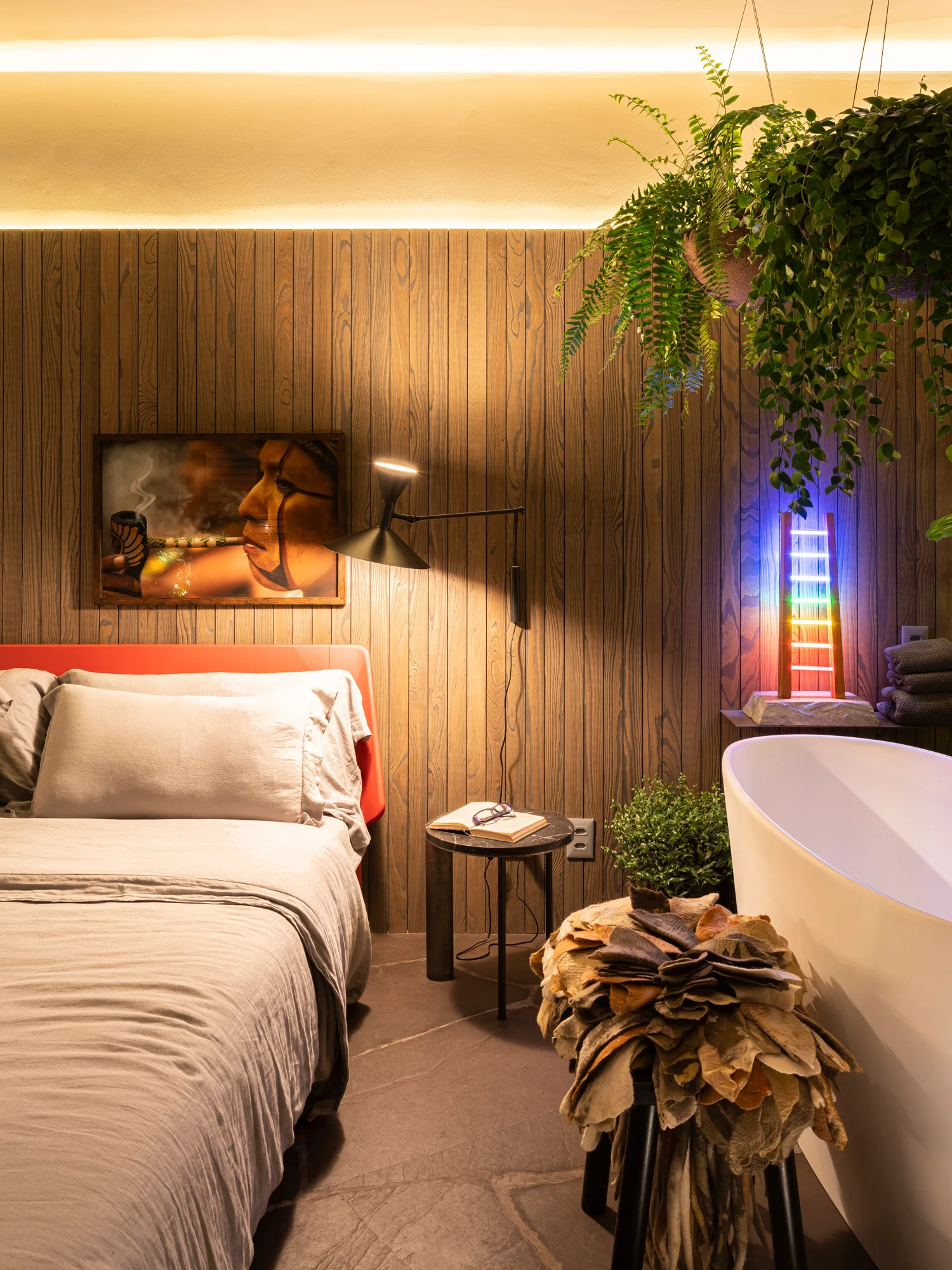 A bedroom with vertical wood wall paneling, a bed with a red headboard, a white freestanding bathtub, and glowing cove lighting.