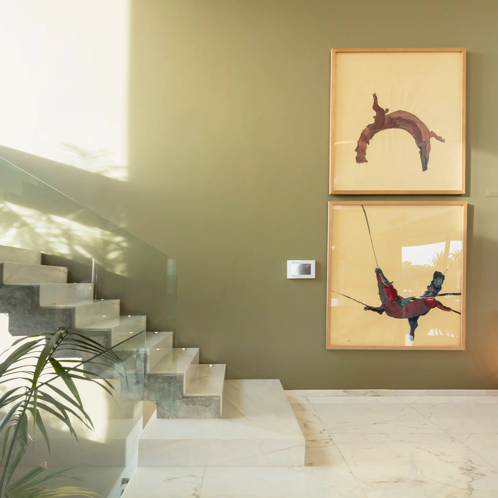 A modern floating marble staircase with a glass railing is set against an olive green wall featuring two stacked, framed art pieces.