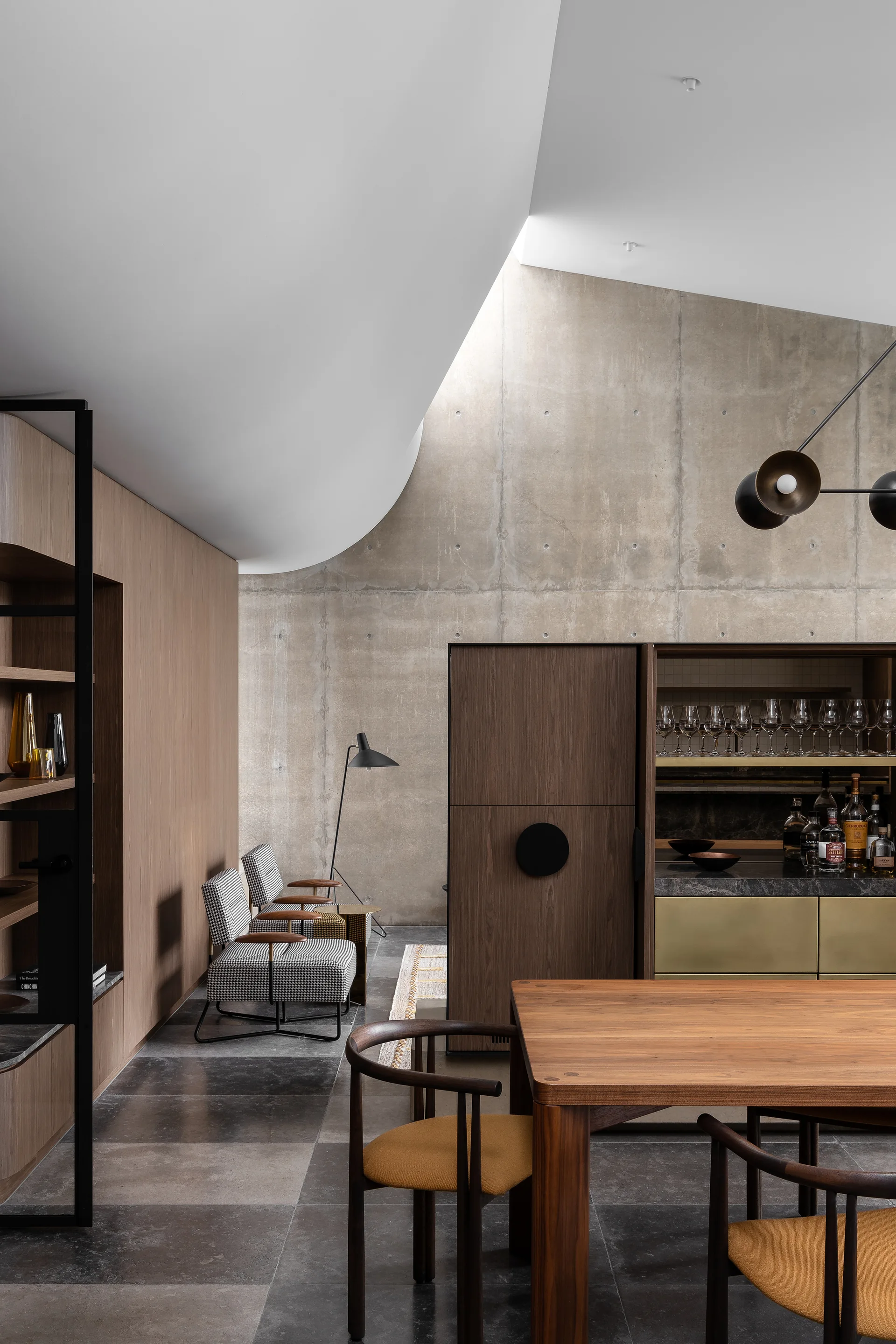 A modern interior dining space features a wooden table, a wood bar cabinet, and two houndstooth armchairs against an exposed concrete wall.