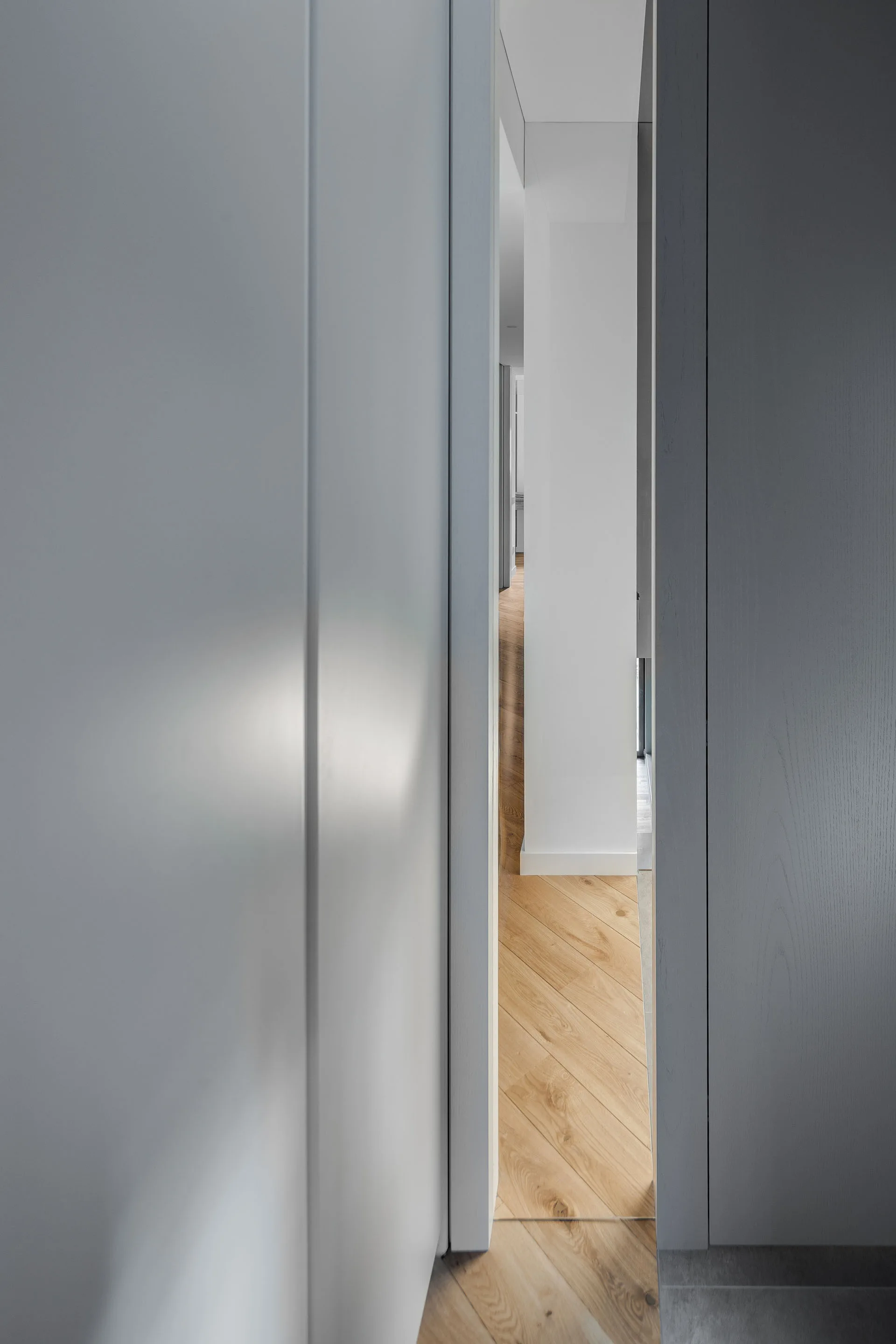 A narrow view through light grey minimalist door panels reveals a bright hallway with herringbone wood flooring and white walls.