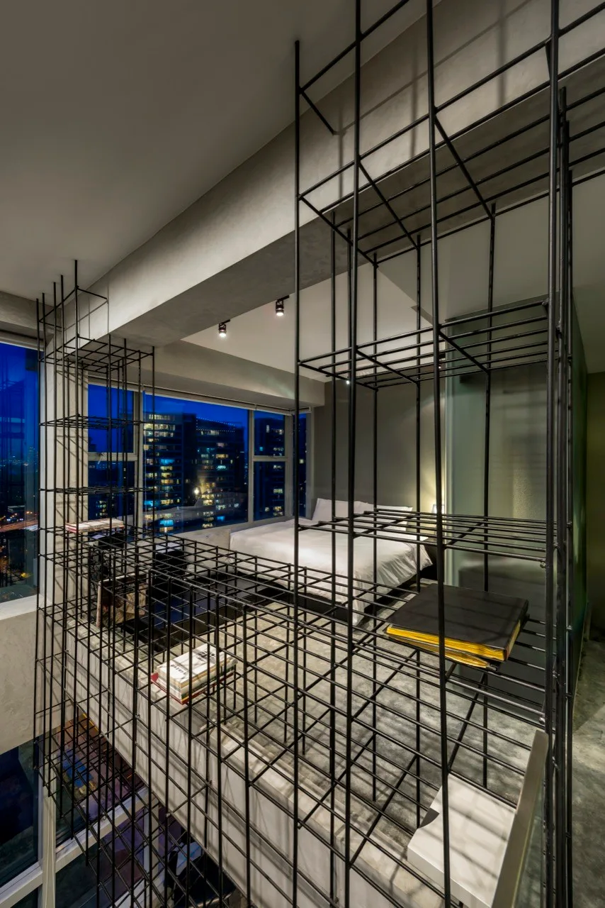 A loft bedroom features a black metal grid shelving unit acting as a barrier beside a bed with white sheets and a large window.