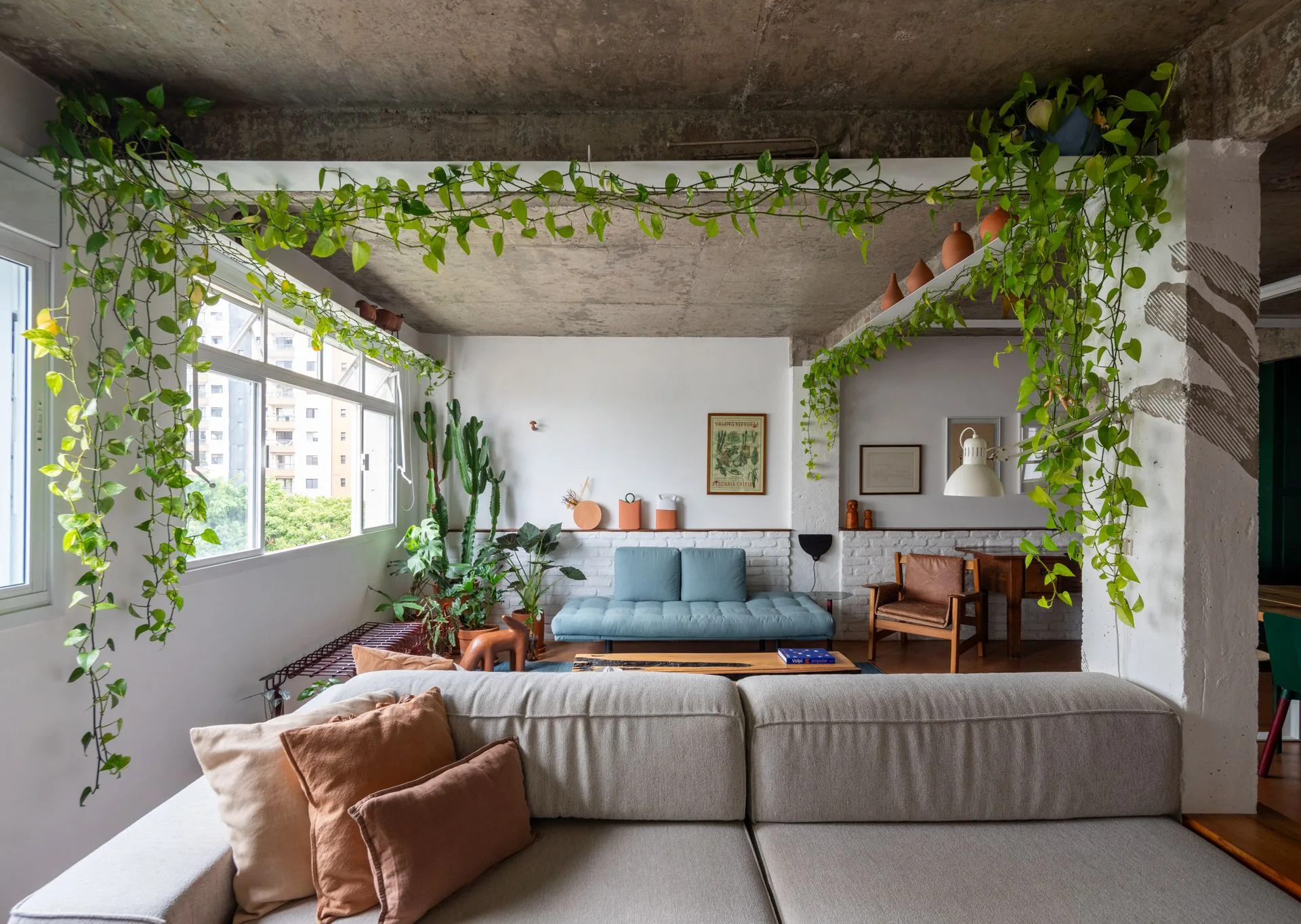 A living room features a large beige sofa, a blue daybed, exposed concrete ceilings, and green vines hanging from overhead beams.