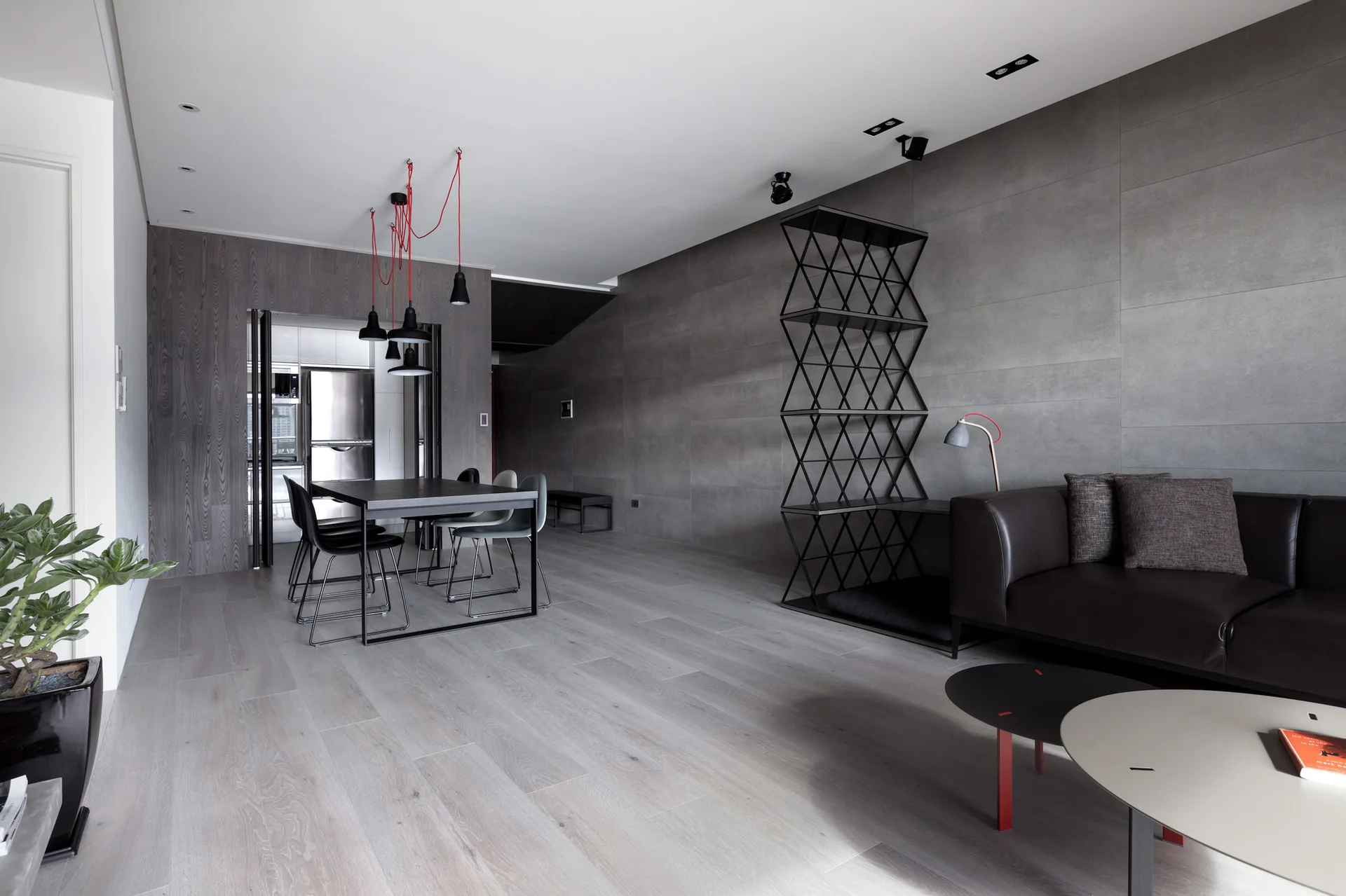 A modern open-plan living room with a grey tiled wall, a geometric black metal shelving unit, and a dark leather sofa.