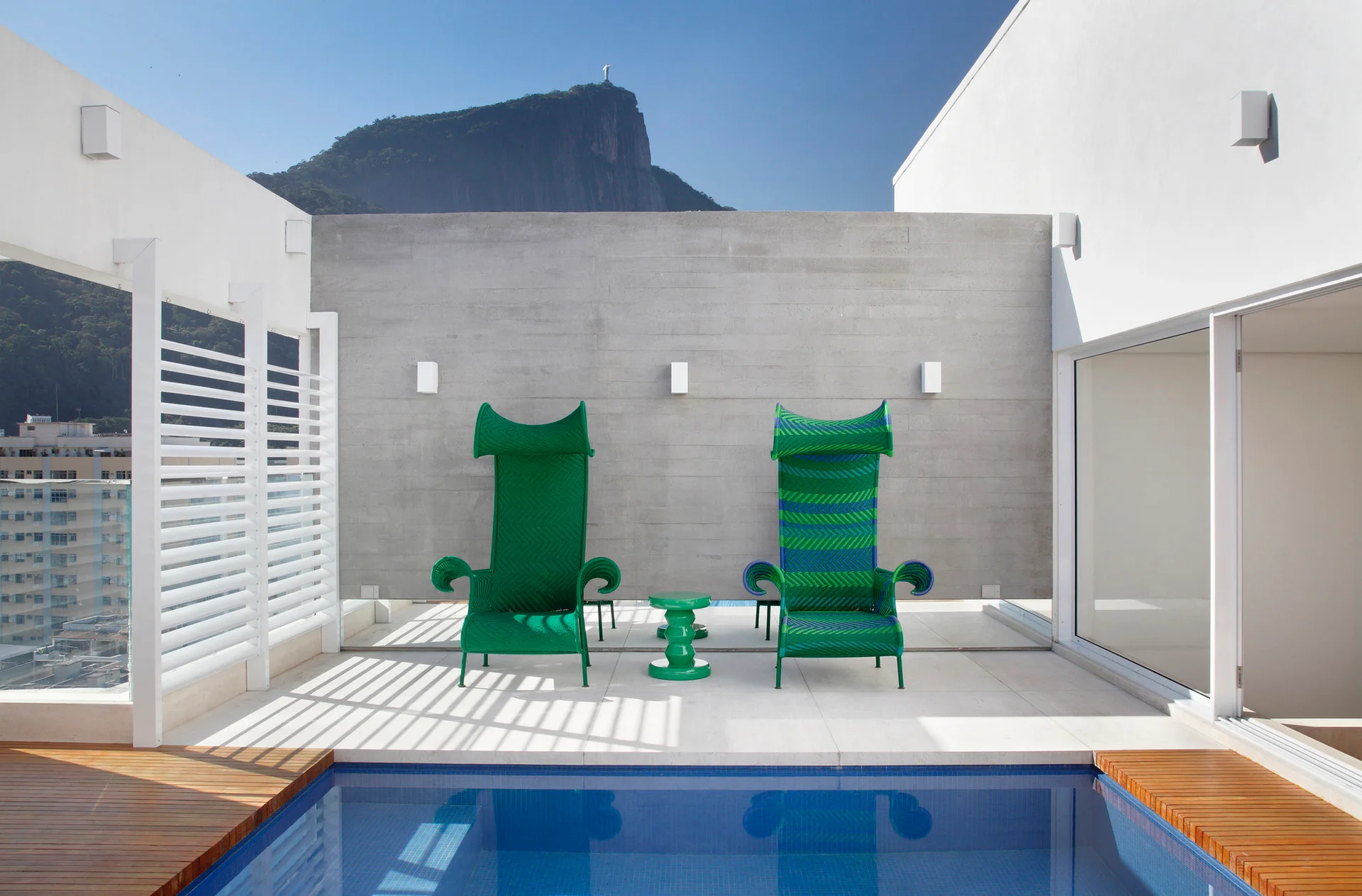 A modern outdoor pool patio features two tall green woven armchairs on white tiling set against a concrete wall under a blue sky.