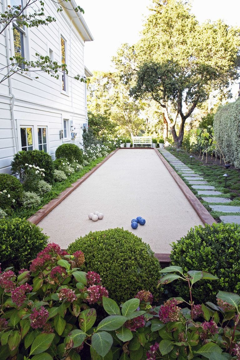 A long sandy bocce court sits in a backyard next to a white-sided house, flanked by manicured boxwood hedges and a stone stepping path.