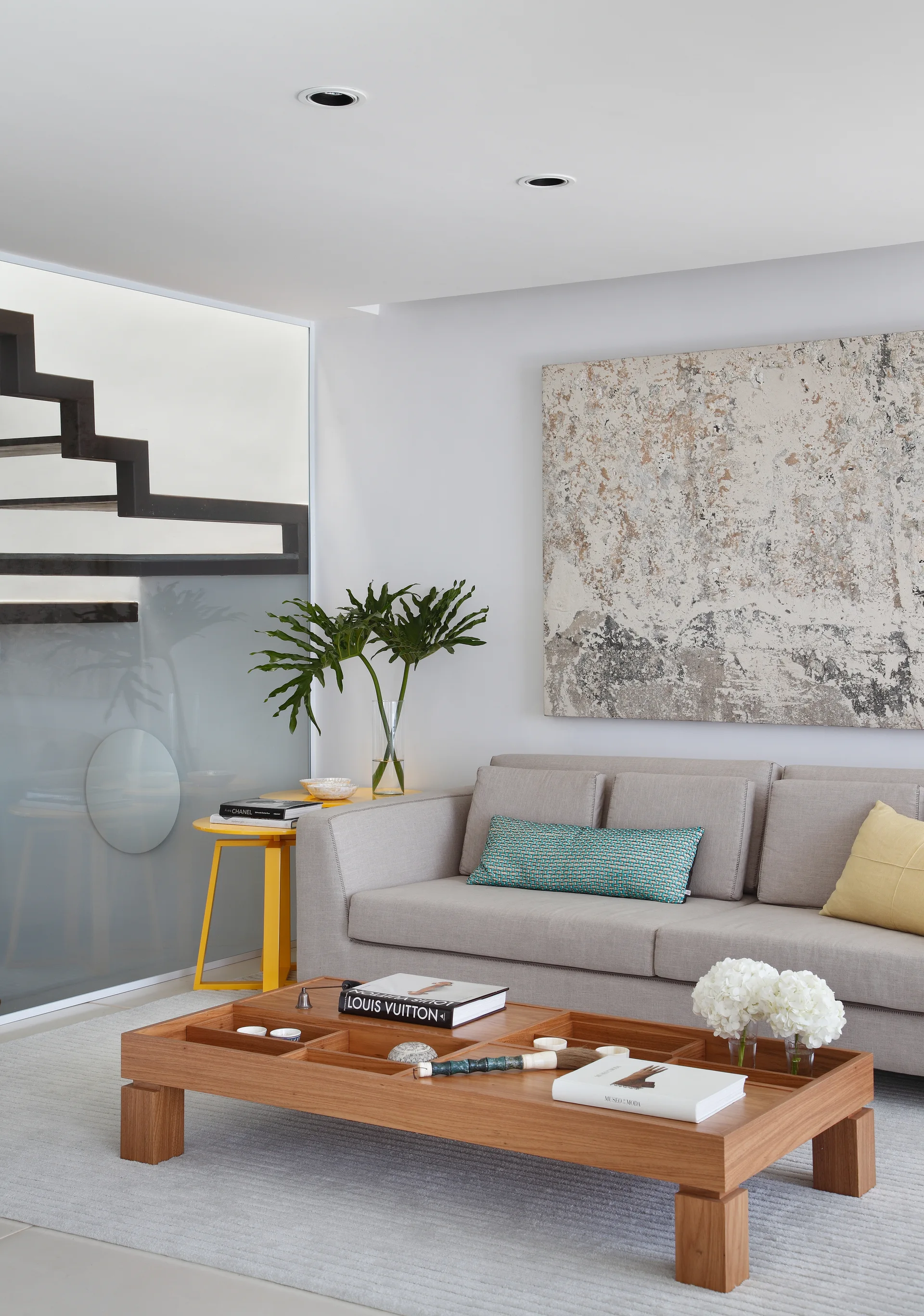 A contemporary living room featuring a grey sofa, a wooden coffee table with built-in compartments, and a staircase behind a frosted glass partition.