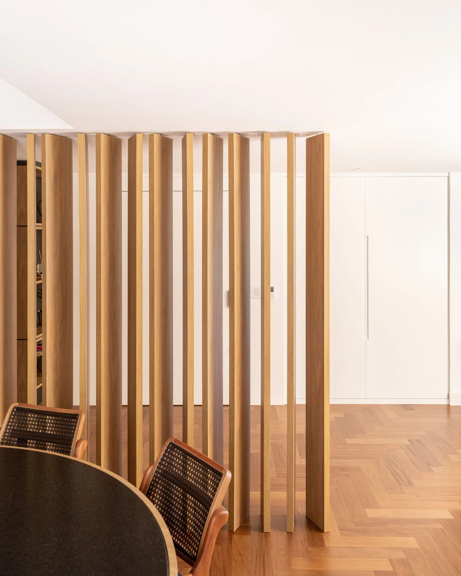 A modern dining area features a vertical wood slat room divider, herringbone parquet flooring, and two caned-back chairs at a dark round table.