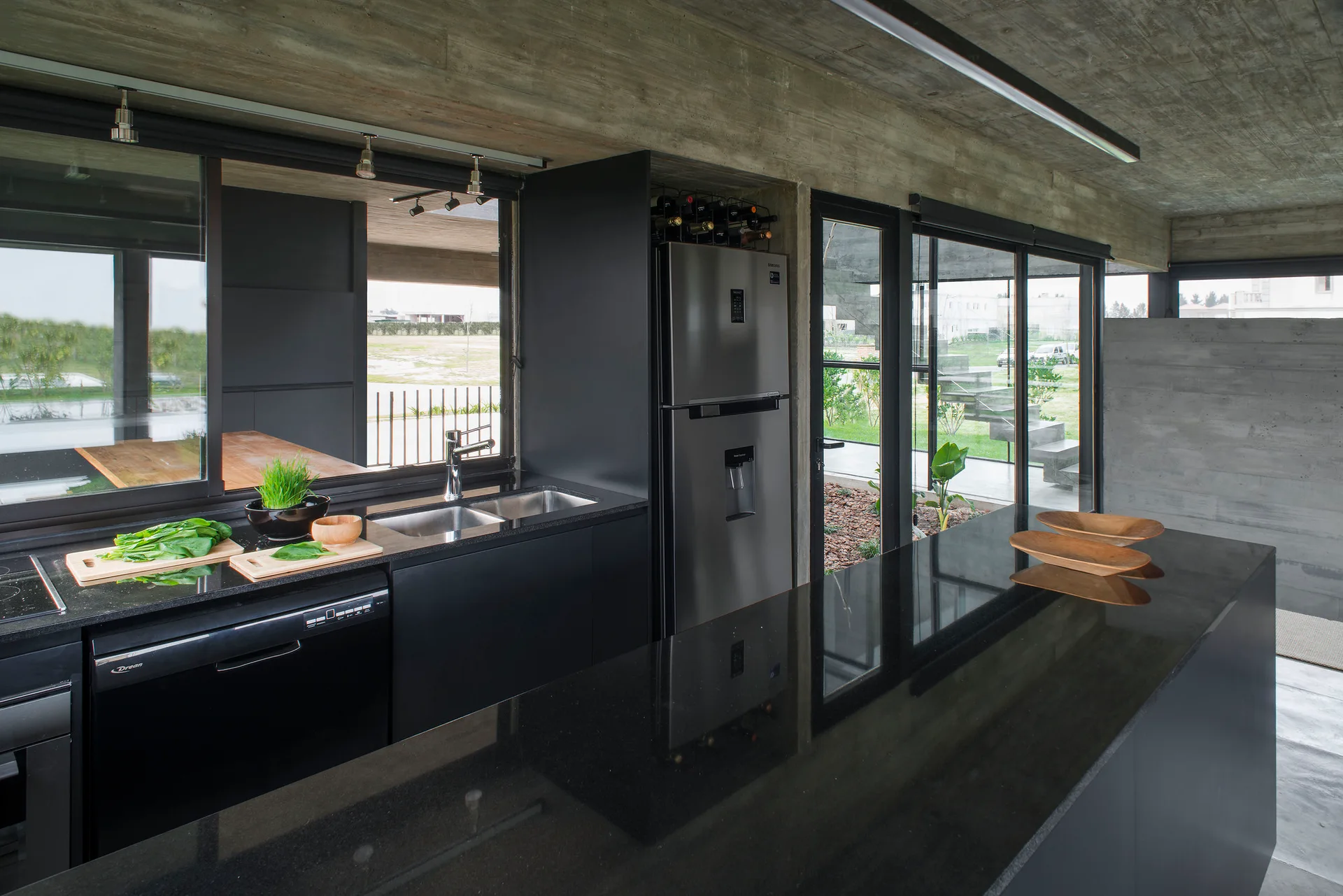 A modern kitchen featuring sleek black lower cabinets, a dark stone island countertop, and a textured, board-formed concrete ceiling.