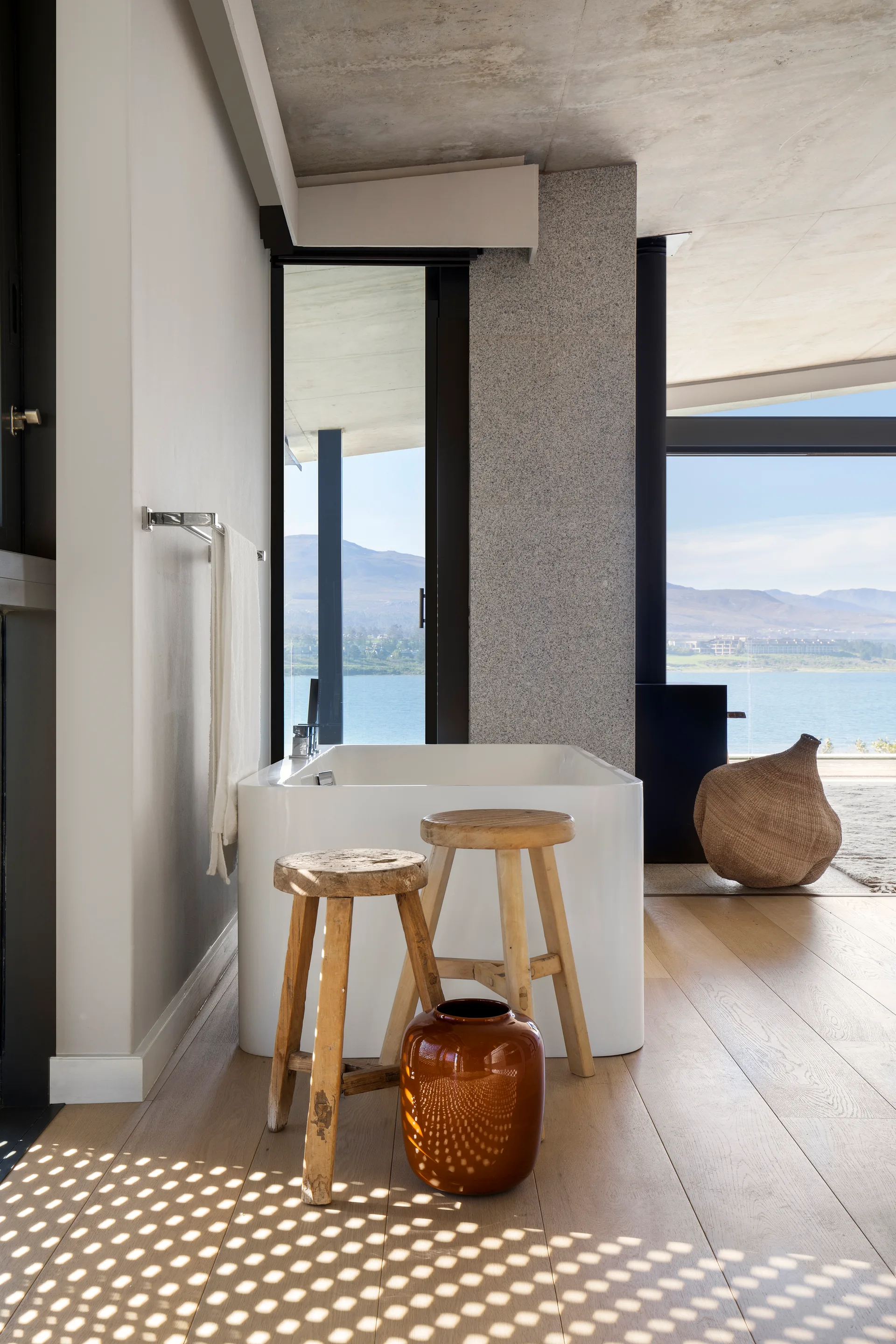 A white freestanding bathtub sits on light wood floors next to two rustic wooden stools and a brown ceramic vase, with a lake view in the background.