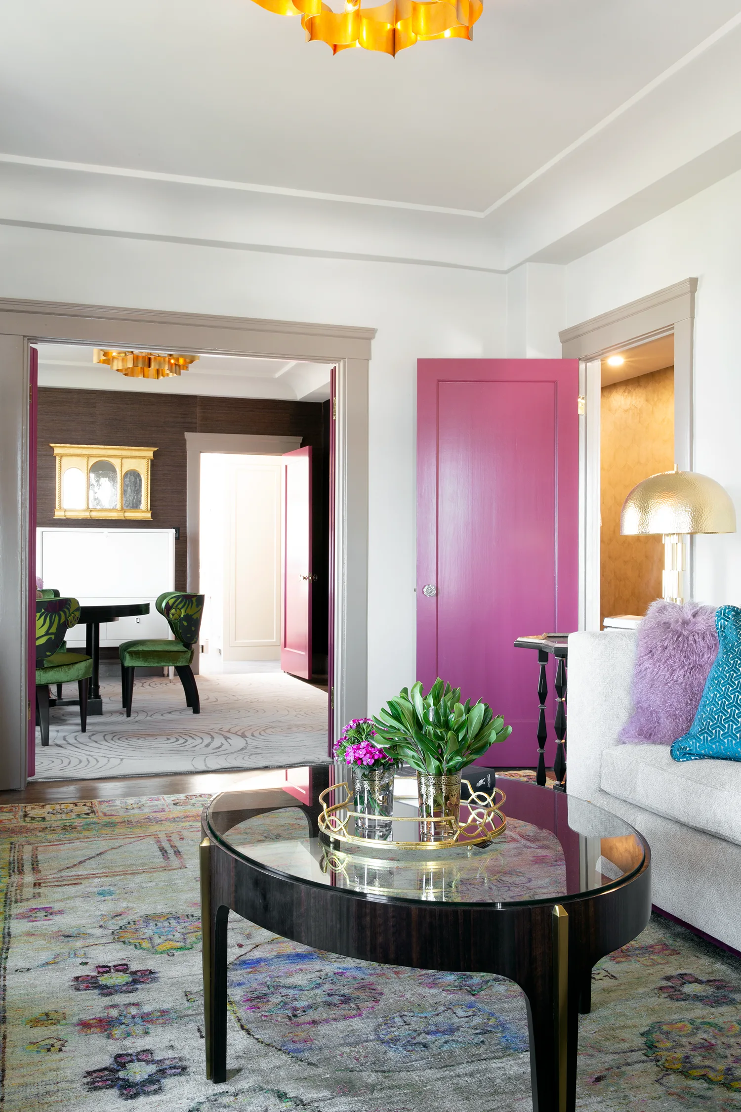 A colorful living room features a bright magenta door, a glass coffee table, a patterned area rug, and a gold dome lamp.