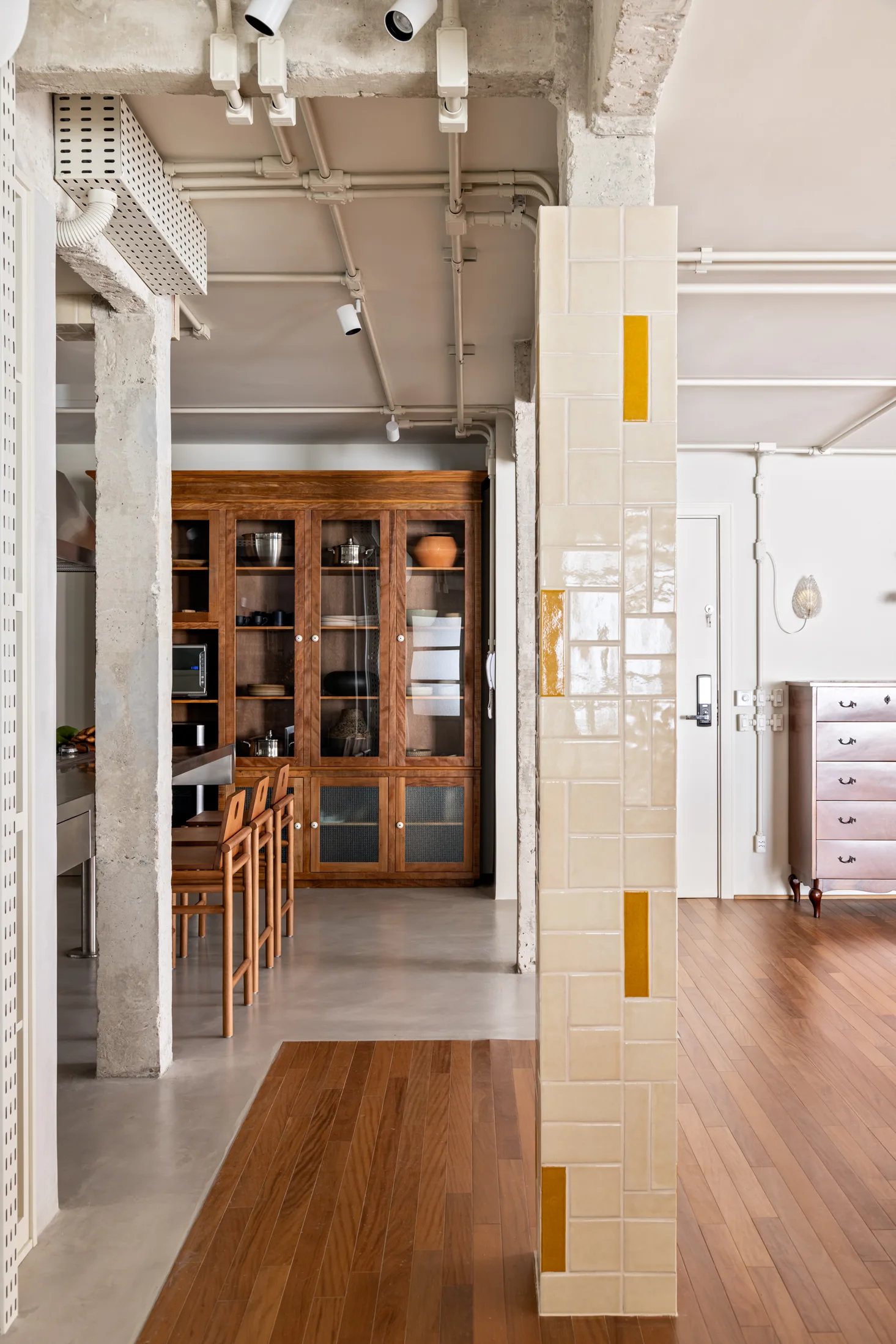 An open-concept kitchen and entryway featuring an industrial concrete ceiling, a wood hutch, and a central pillar wrapped in glossy beige and yellow tiles.
