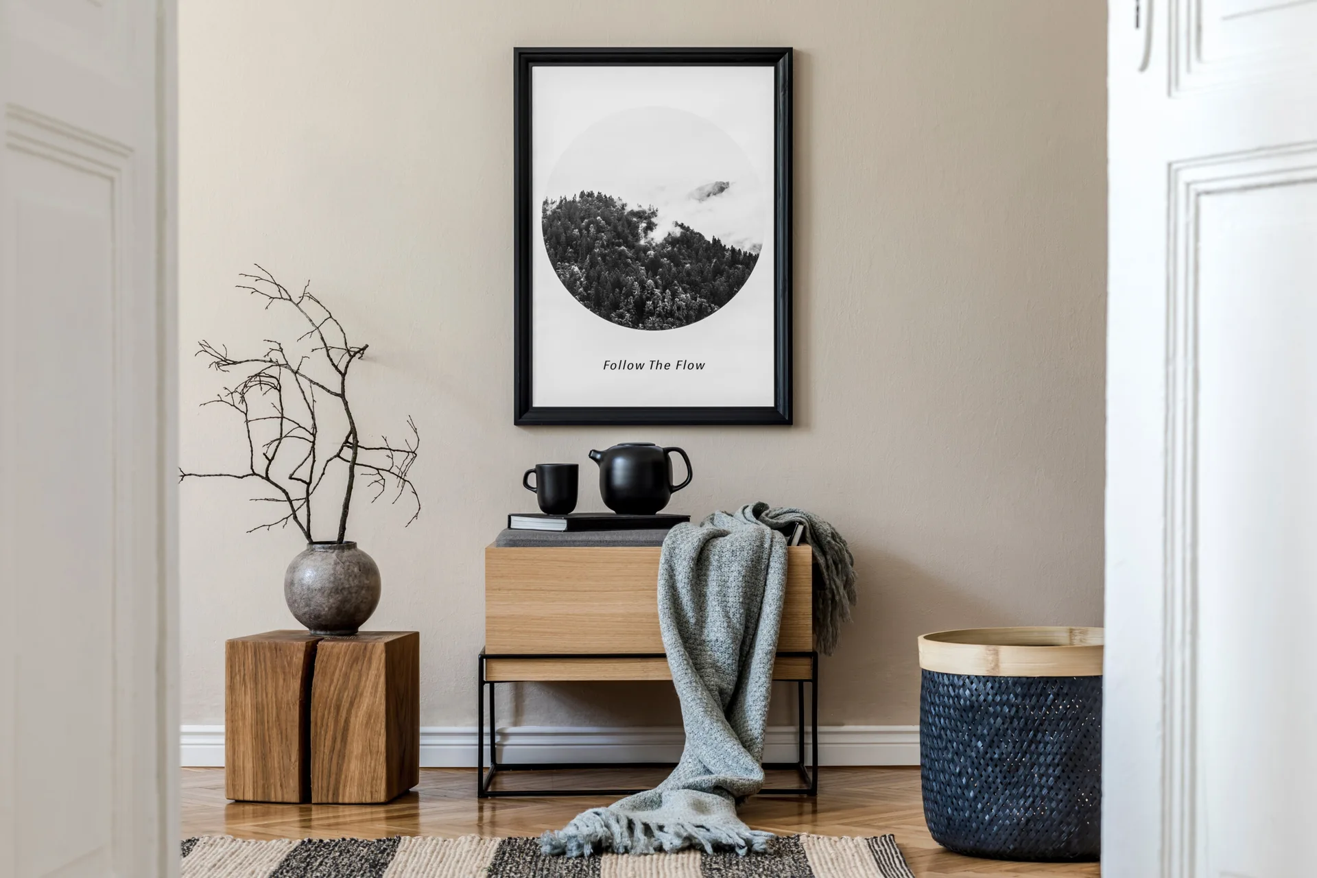 A wooden console bench with a draped grey throw blanket sits beneath framed wall art next to a rustic wood block stool holding dry branches.