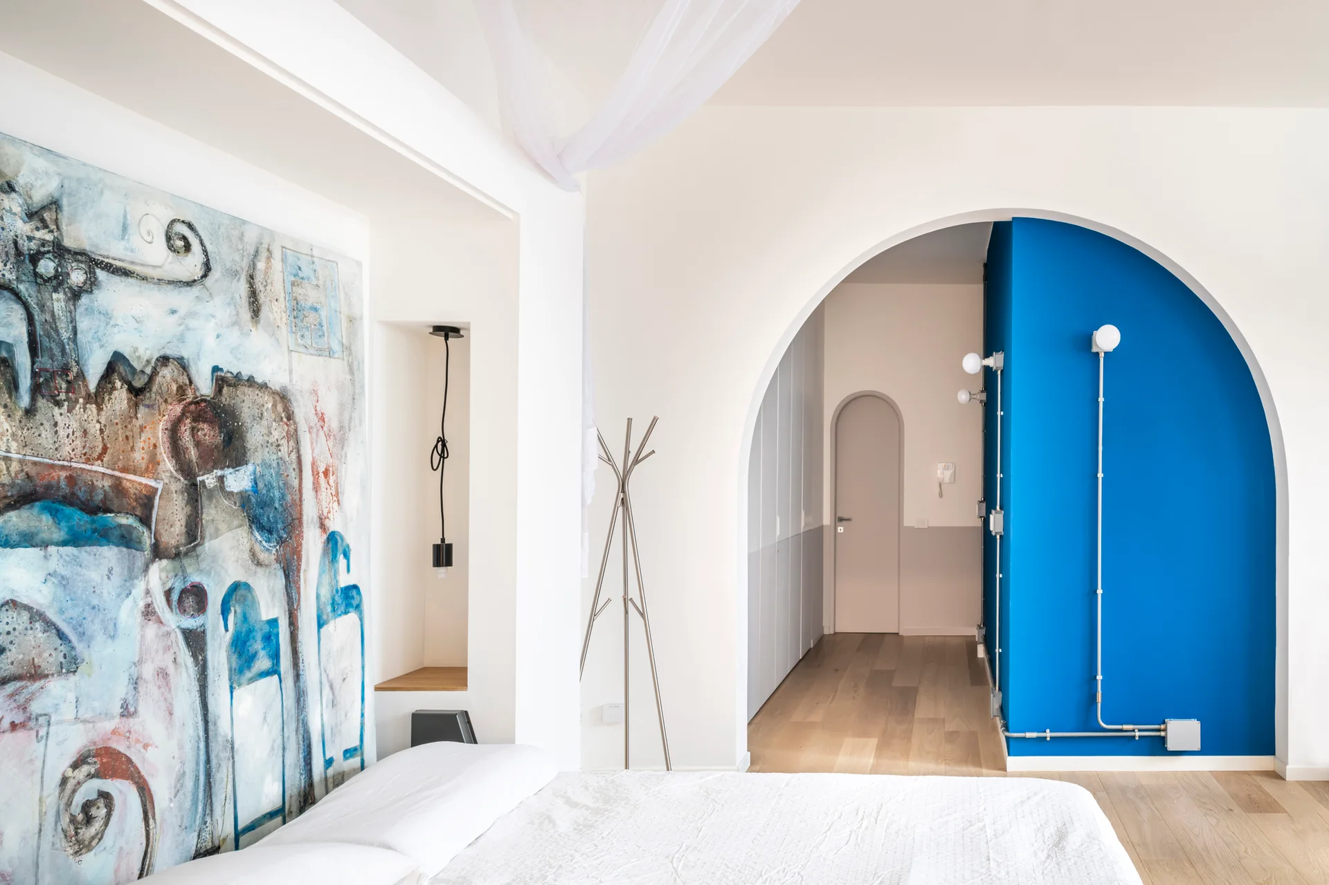 A minimalist white bedroom featuring a large abstract painting, a recessed wall niche, and an archway leading to a bright blue hallway wall.