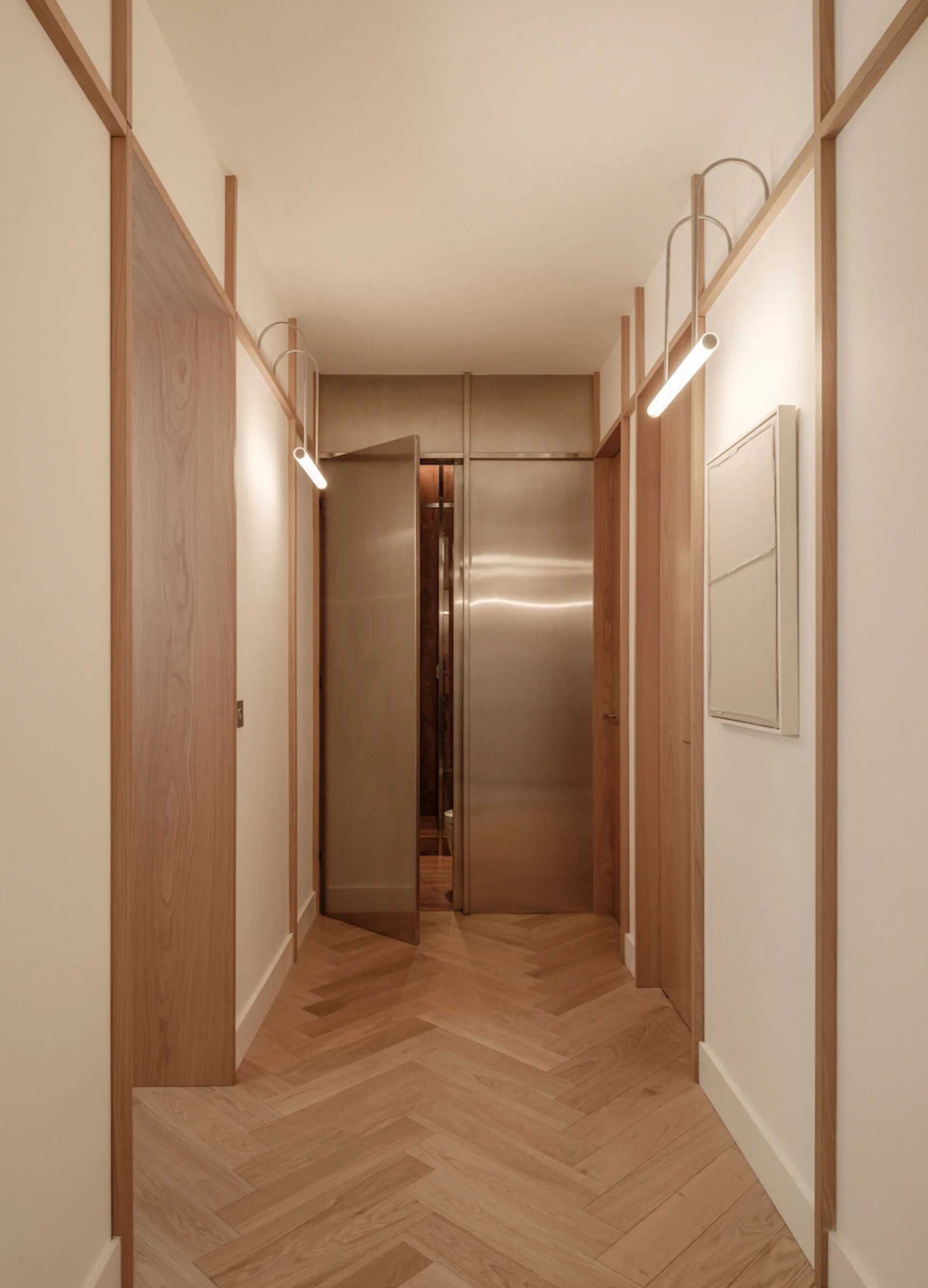 A narrow hallway features light oak herringbone floors, white walls with vertical wood trim, curved tubular wall lights, and brushed silver metal doors.