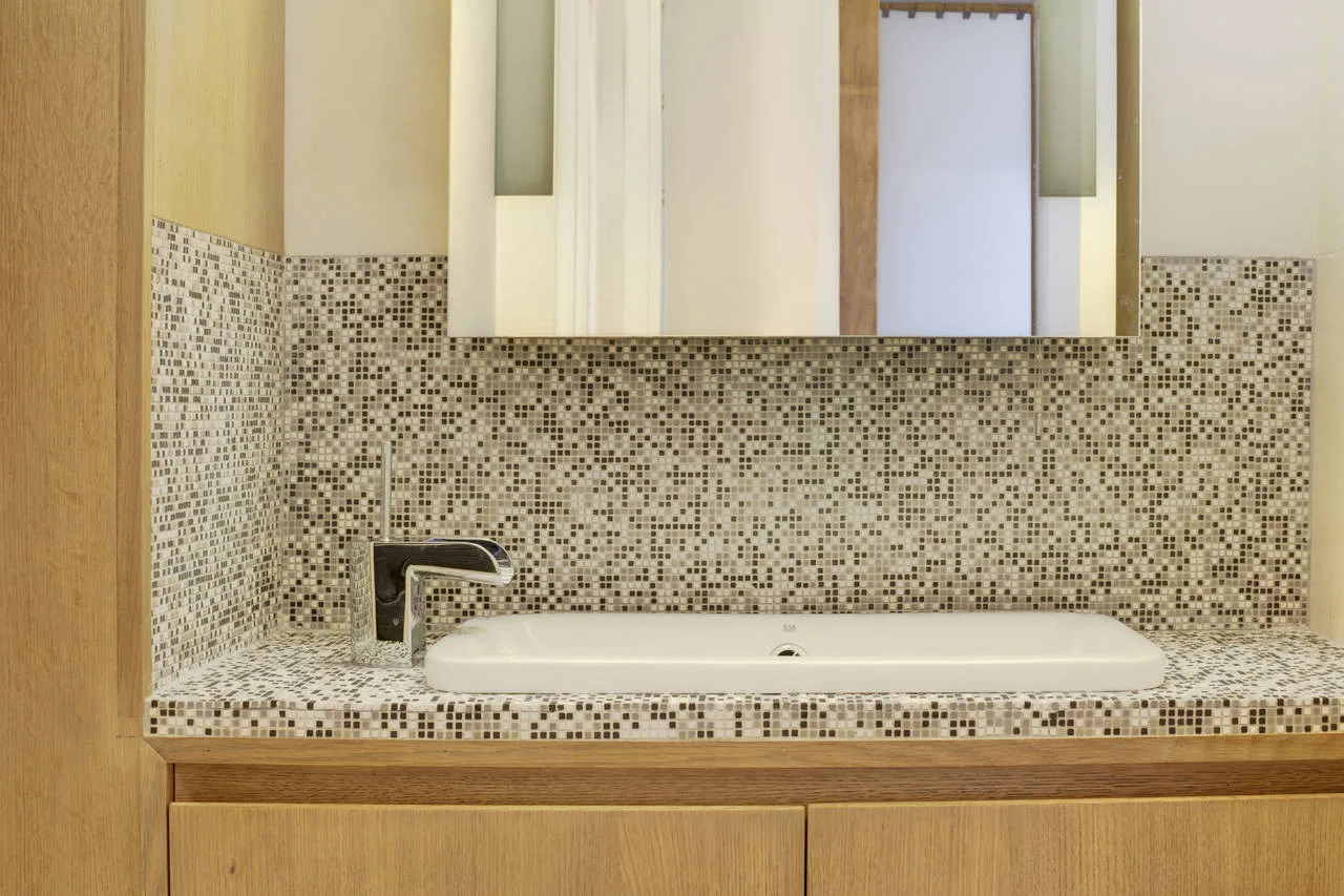 A modern bathroom vanity featuring a white rectangular sink with a chrome faucet set against a brown and beige mosaic tile countertop and wall.