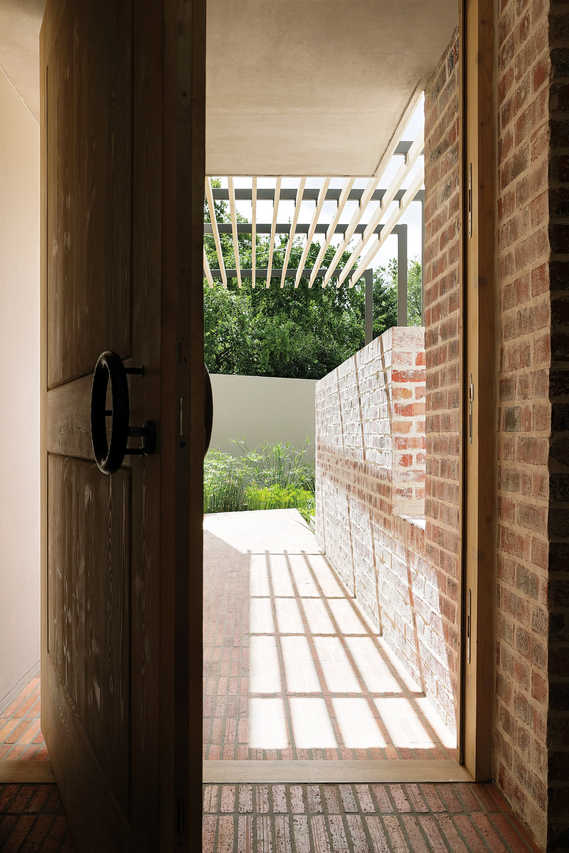 An open wooden entryway door looks out onto a brick patio under a slatted timber pergola casting linear shadows.