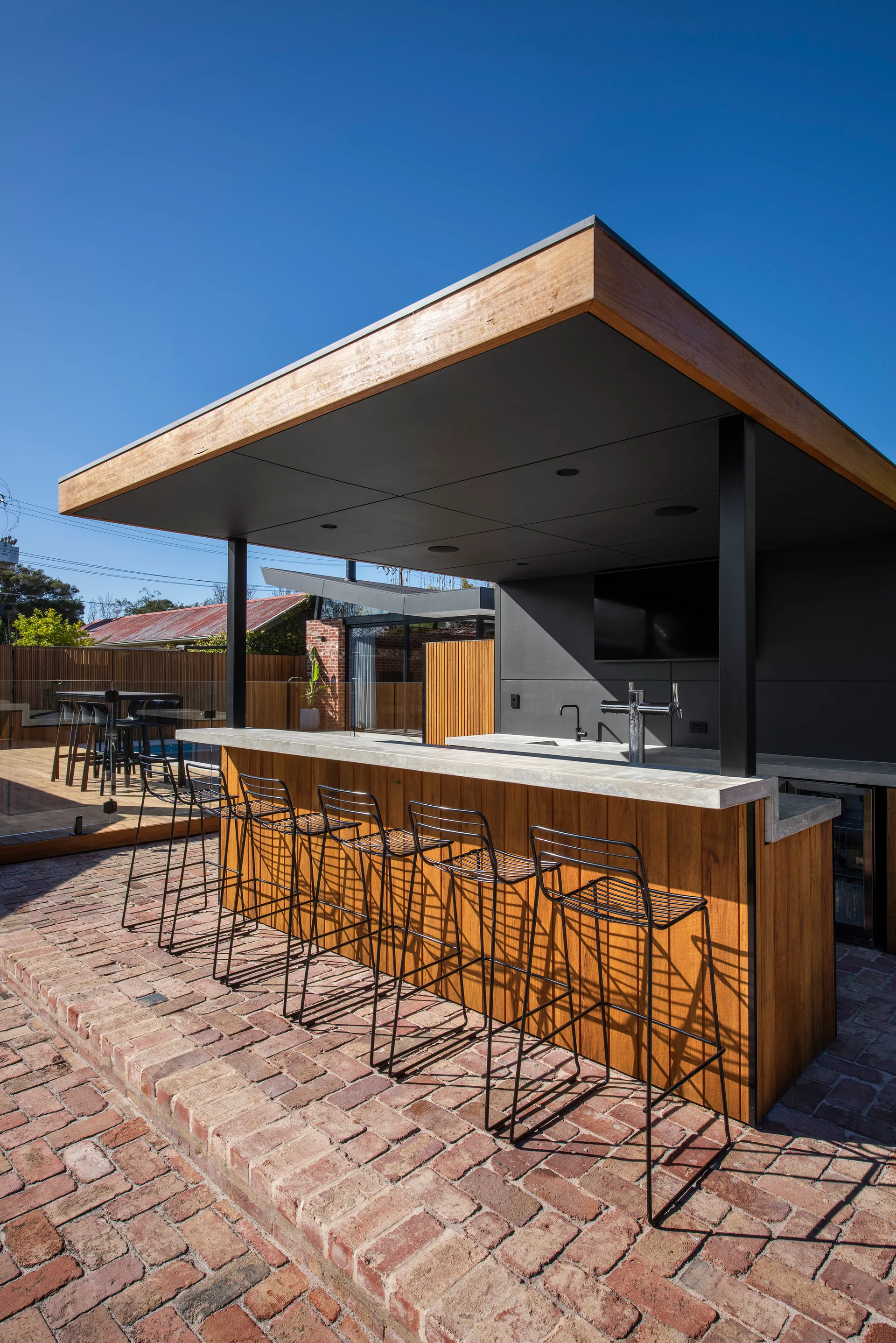 A covered outdoor bar on a brick patio featuring a wood-clad counter, a concrete top, and black wire barstools.