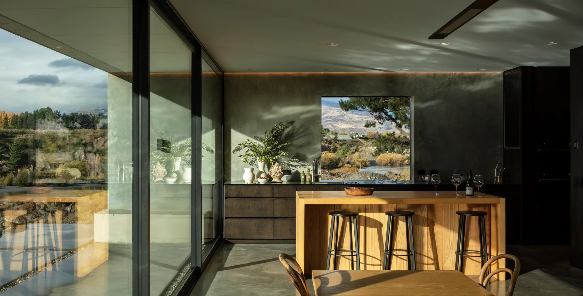 A modern kitchen features a warm wood island, concrete walls, dark lower cabinets, and large glass windows displaying a rugged landscape.