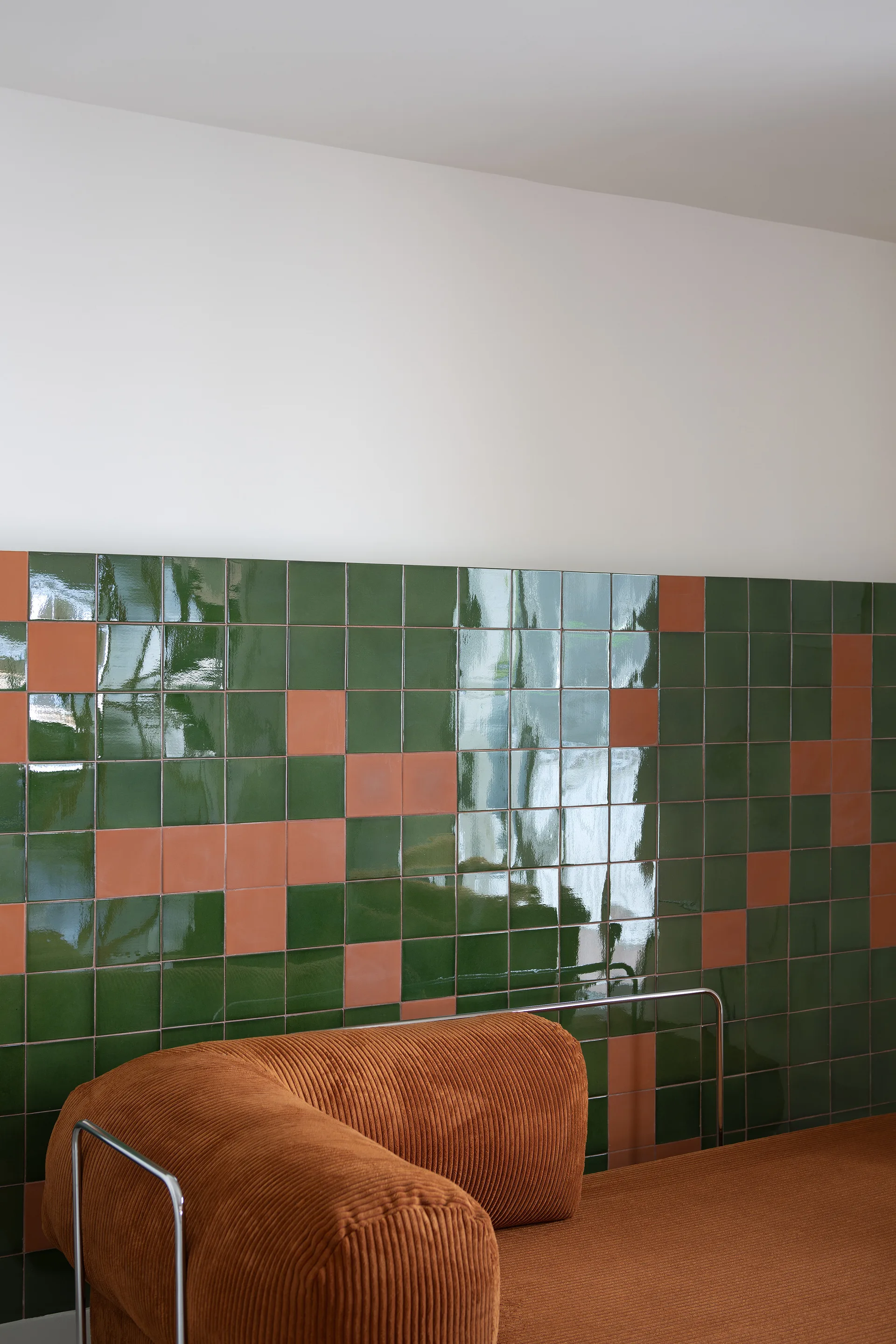 An orange corduroy sofa stands against a wall clad in glossy green square tiles with a scattering of orange accent tiles.