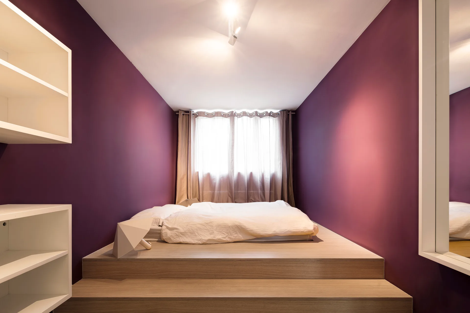 A minimalist bedroom featuring deep purple walls, a raised light wood platform bed with white sheets, and white built-in shelving.