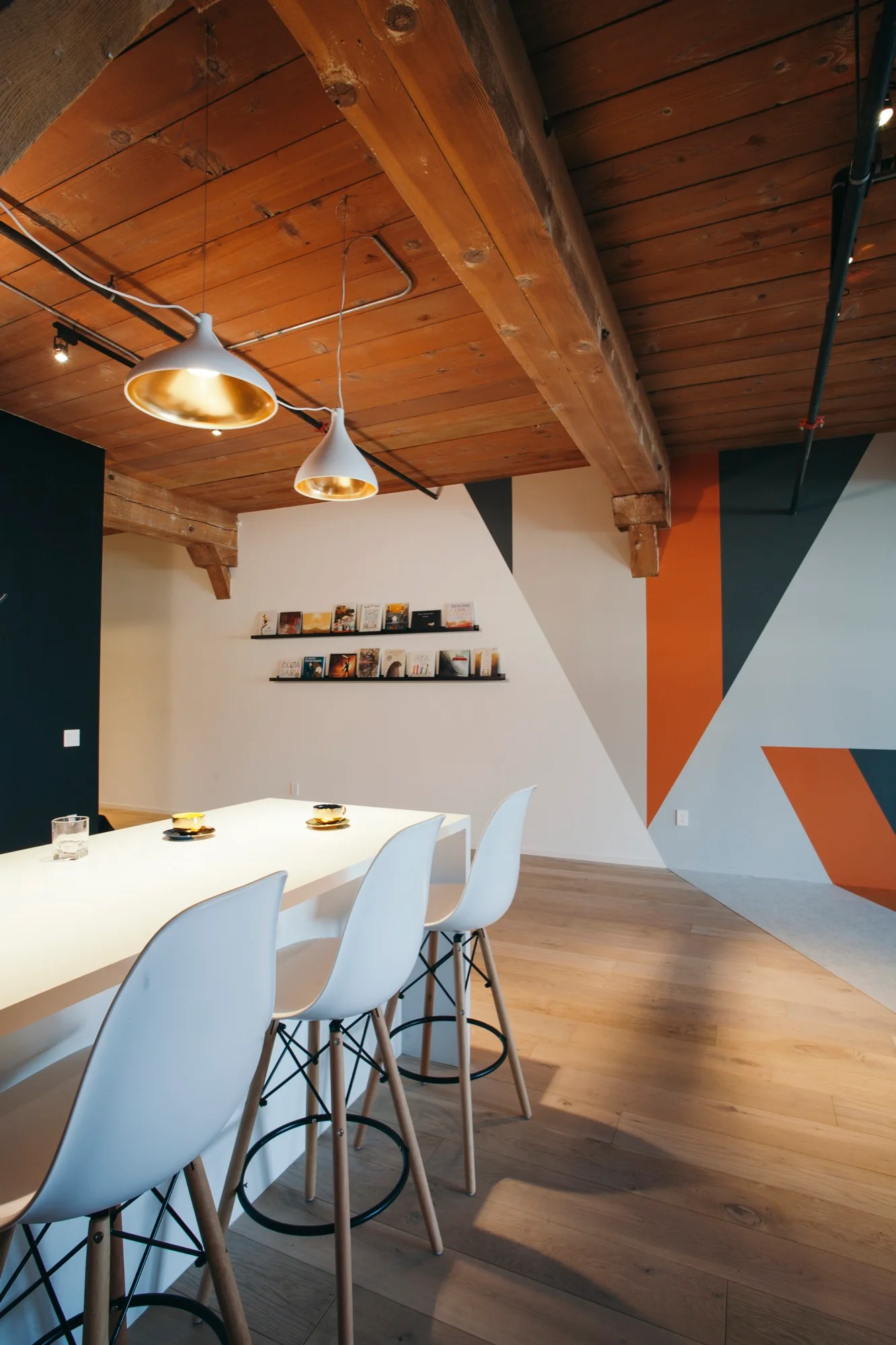 A modern dining space features white bar stools at a long white counter under an exposed timber ceiling and a bold geometric wall mural.