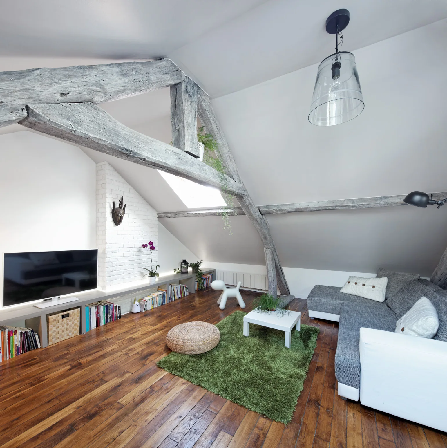 An attic living room featuring a grey sectional sofa, a green shag rug, a low bookshelf, and prominent rustic wood beams supporting a slanted ceiling.