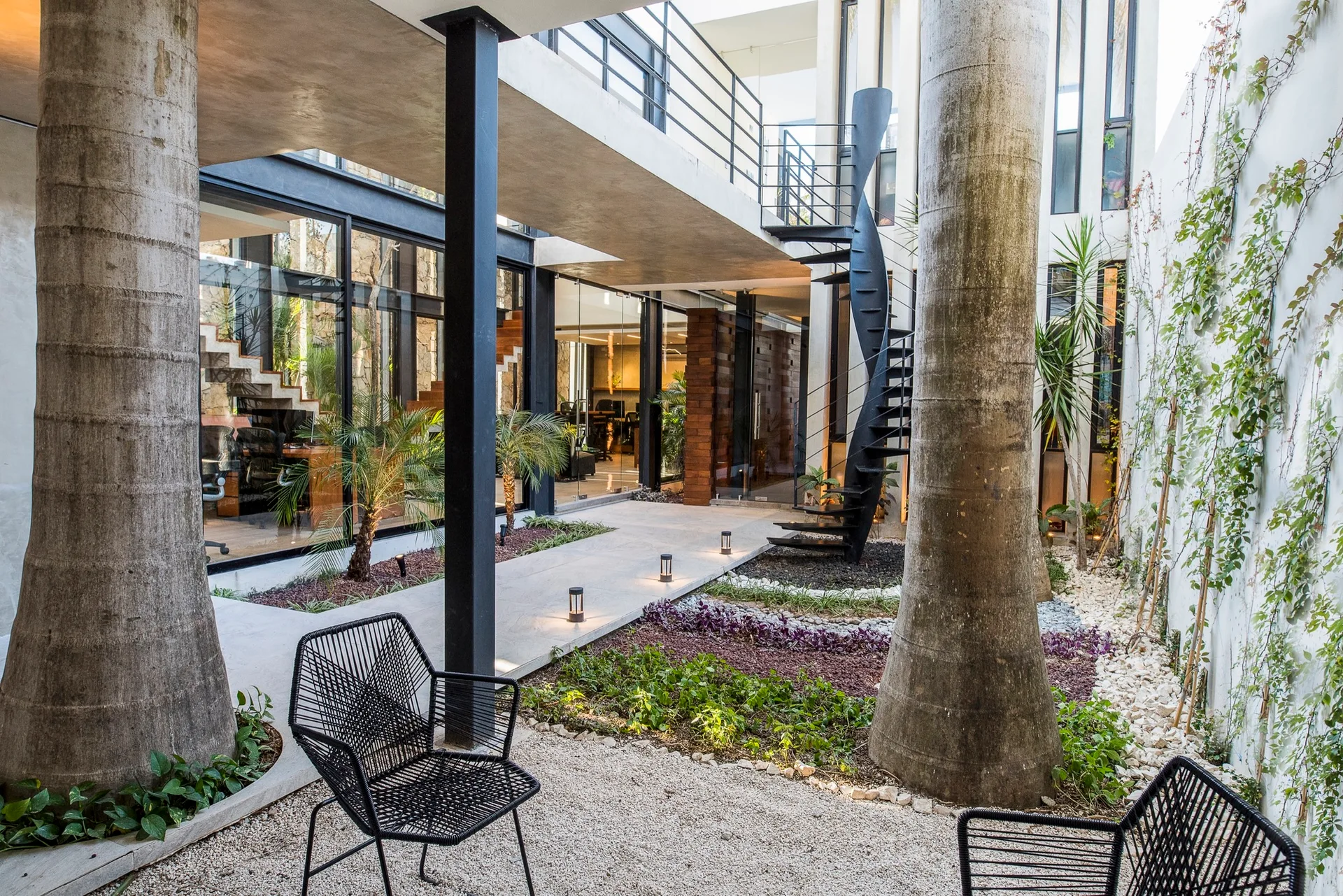 An open-air courtyard features a concrete walkway, a black spiral staircase, gravel landscaping, and two black wire chairs.