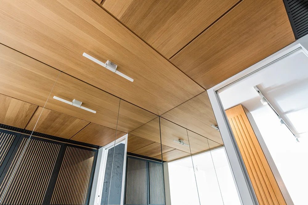 Warm wood-toned ceiling panels meet a large mirrored wall reflecting dark vertical slatted cabinetry and linear light fixtures.