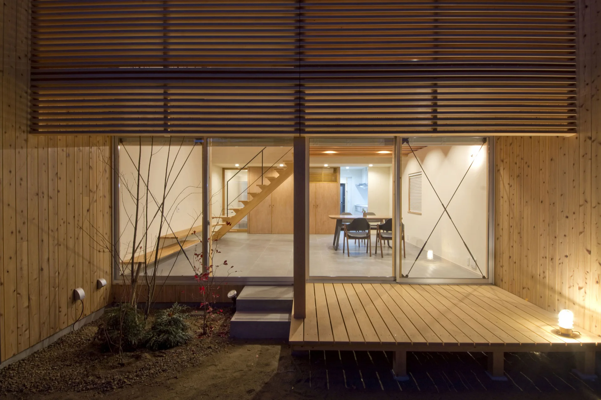 An exterior view of a modern wood-clad home looking through large sliding glass doors into a minimalist interior with a wooden staircase and dining set.