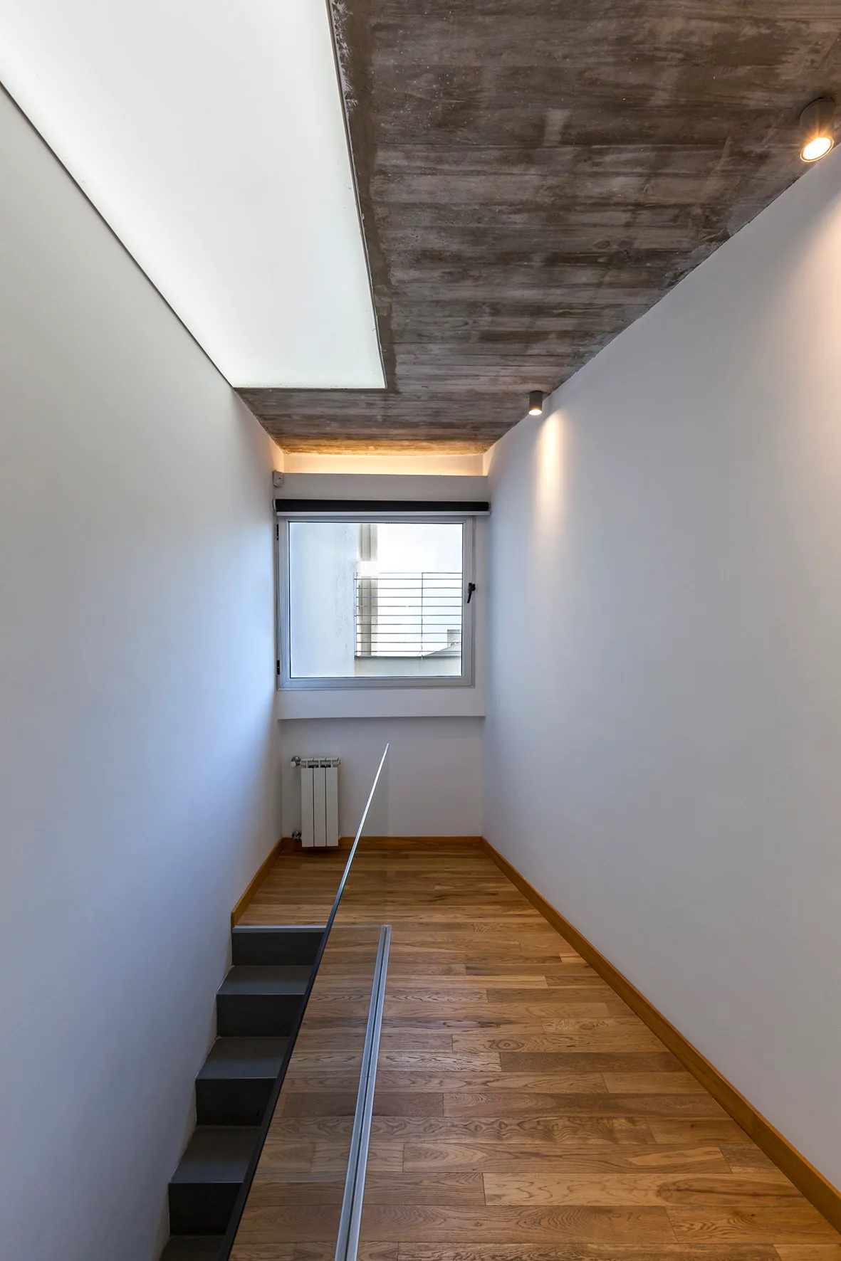A minimalist staircase landing features a raw concrete ceiling, wooden plank flooring, white walls, a glass railing, and a window at the end.