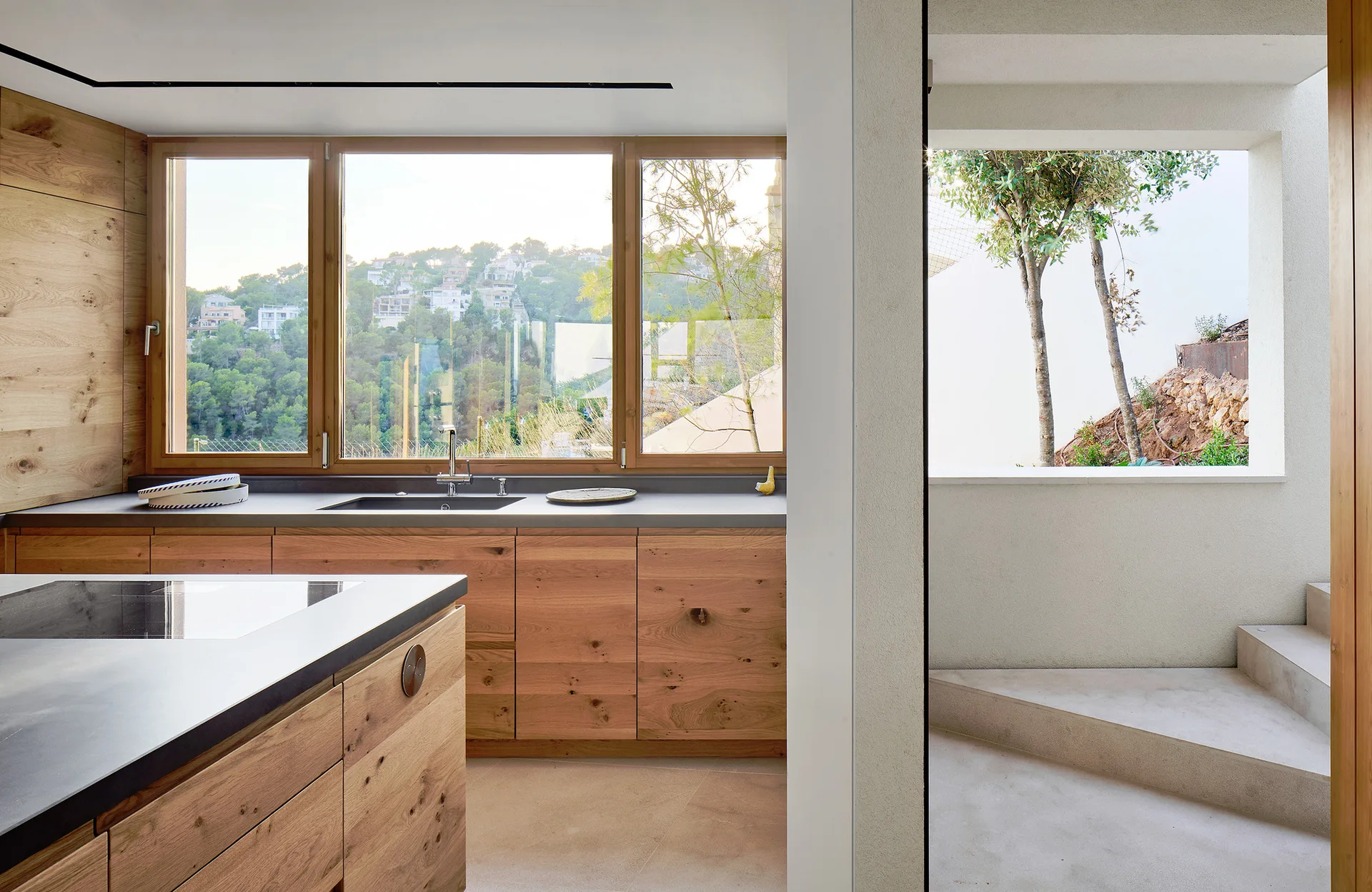 A modern kitchen features warm knotty wood cabinets, dark grey countertops, a wood-framed window, and adjacent concrete steps.